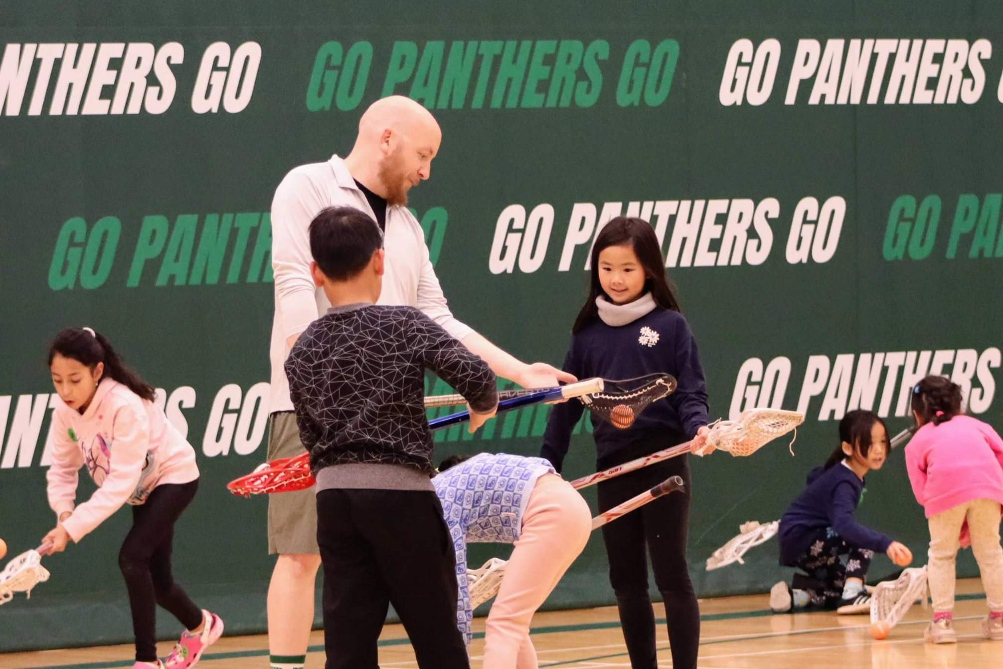 Children practicing lacrosse on a gym floor with a coach, in front of a green background with the words 'Go Panthers' written repeatedly.