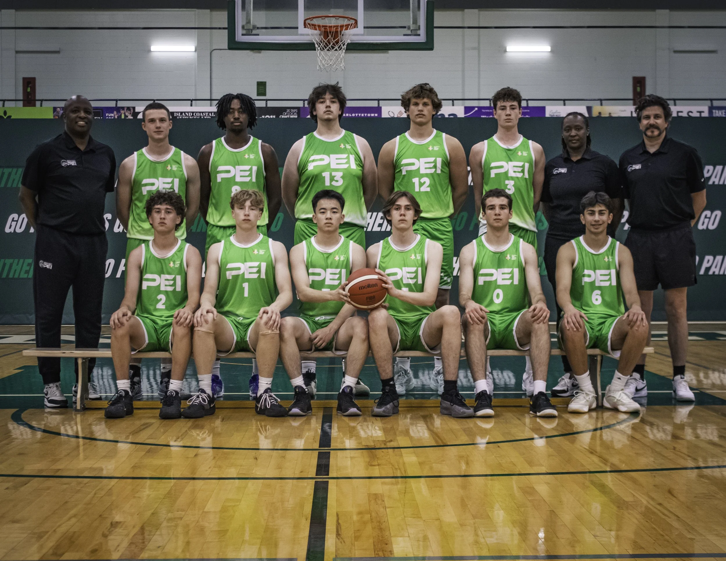 A youth basketball team in green uniforms poses in a gym with their coaches. The team is organized in two rows, with some players sitting and others standing, holding a basketball for the photo.