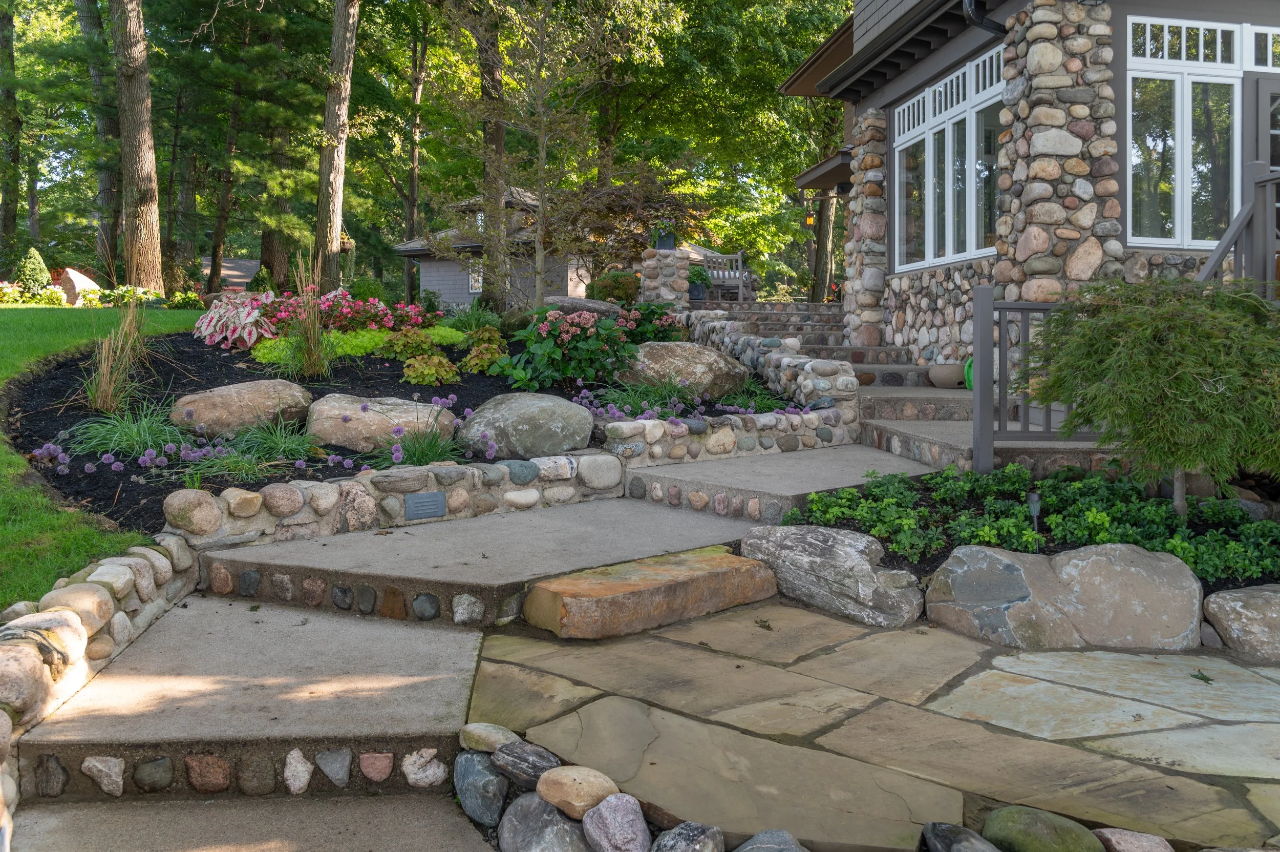 A landscaped front yard with stone pathways, flower beds with pink and purple flowers, large rocks, and a house with stone and large window walls, surrounded by tall trees.