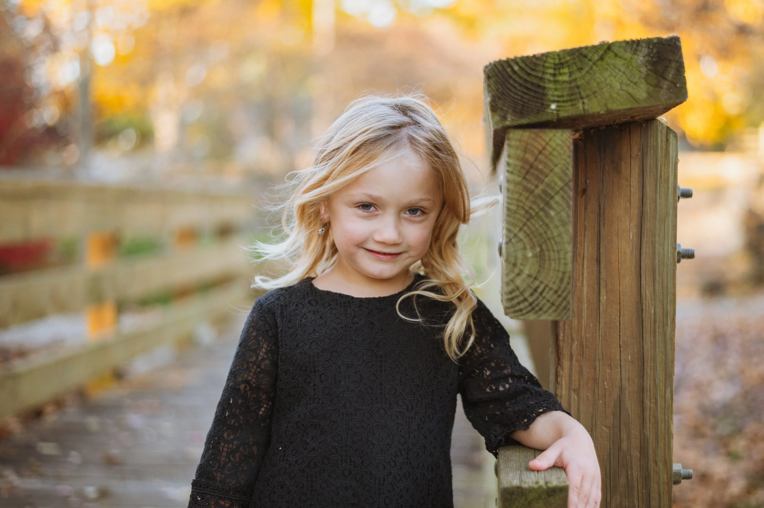 A young girl with blonde hair wearing a black lace dress, standing outdoors on a wooden walkway, leaning on a wooden post during autumn.