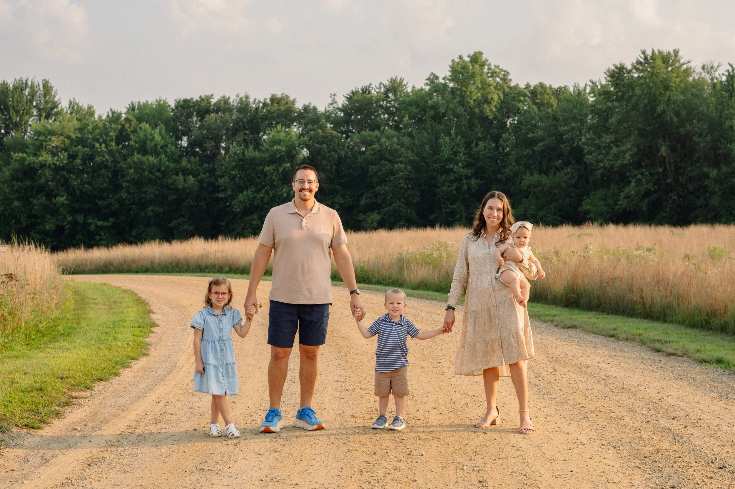 Smiling family of five holding hands on a dirt country road during sunset, with grassy fields and trees in the background