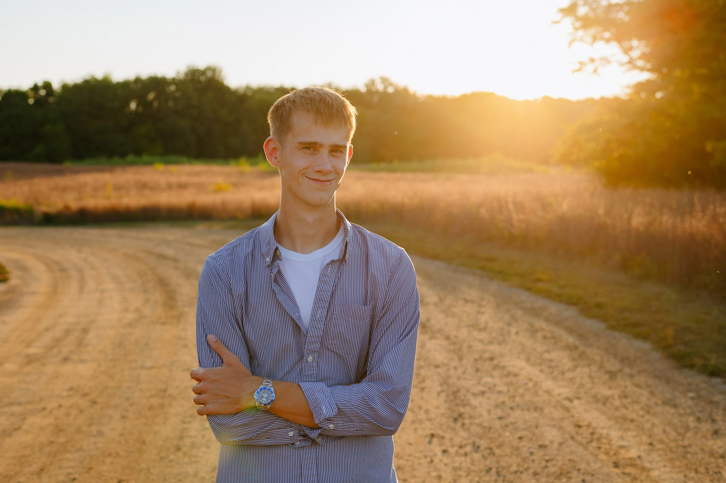 A young man stands on a dirt road during sunset, smiling, with his arms crossed, wearing a striped shirt and a wristwatch.
