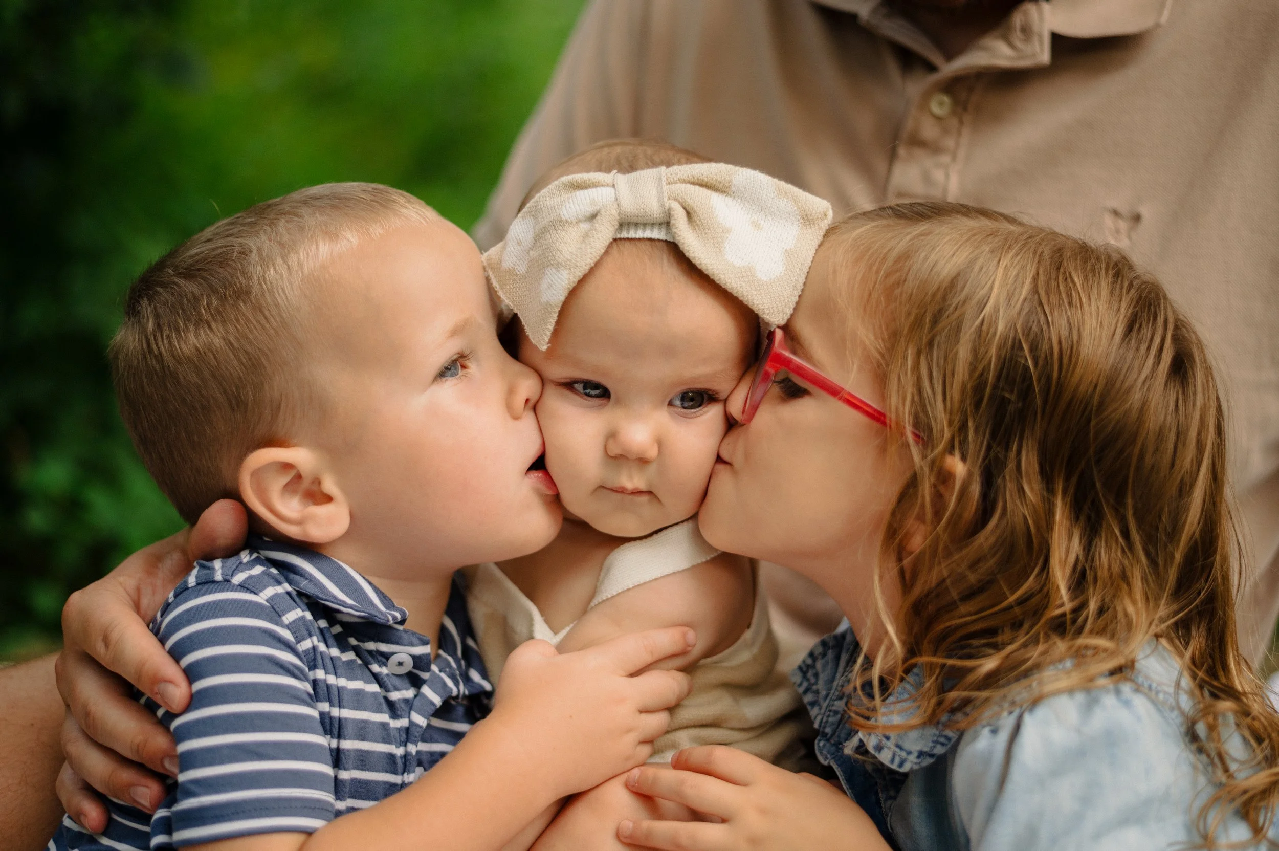 A woman with red hair and glasses kissing a baby on the cheek, surrounded by two young children, outside with green foliage in the background.