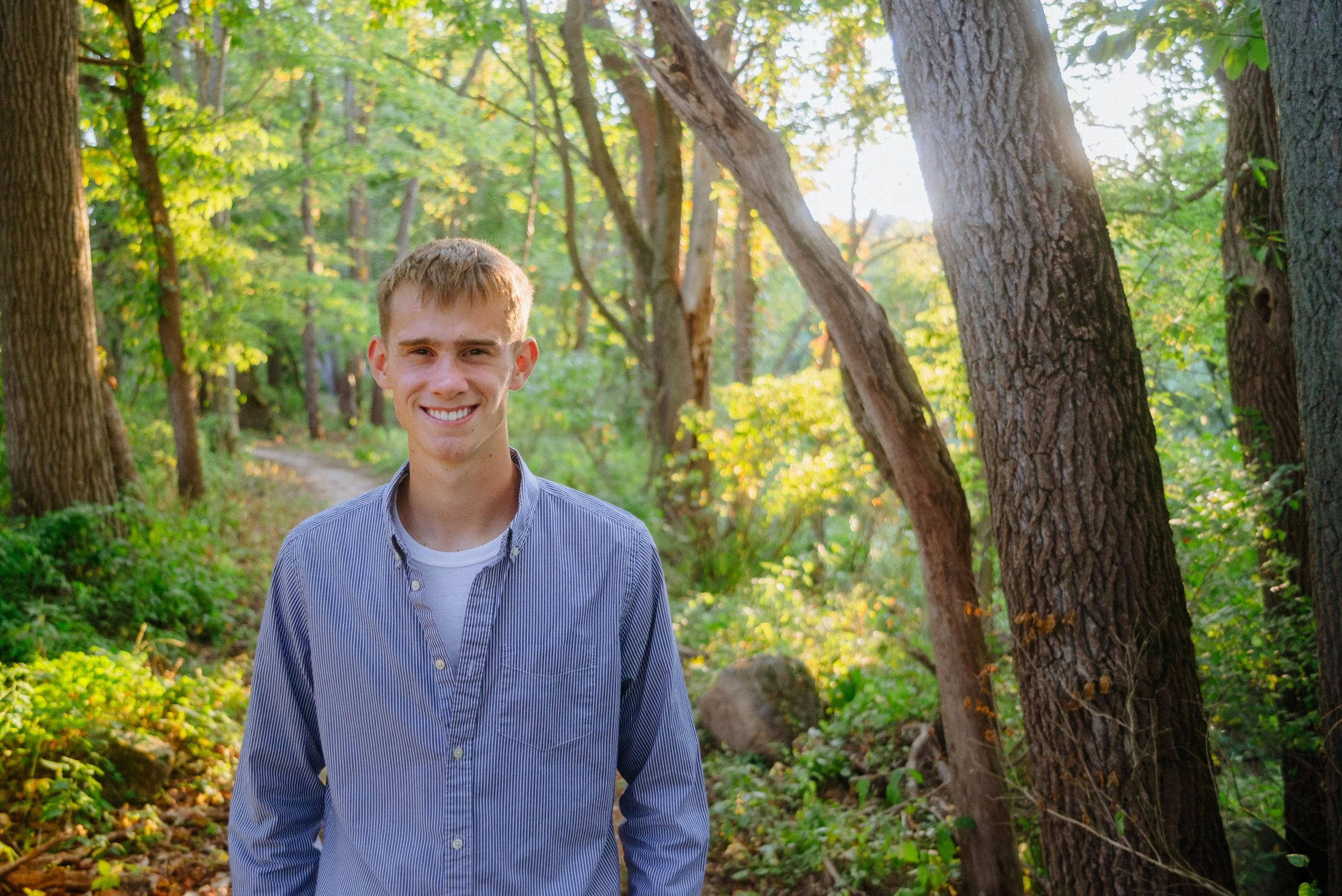 A young man standing on a wooded trail, smiling and wearing a blue striped shirt and white T-shirt, with sunlight filtering through green trees.