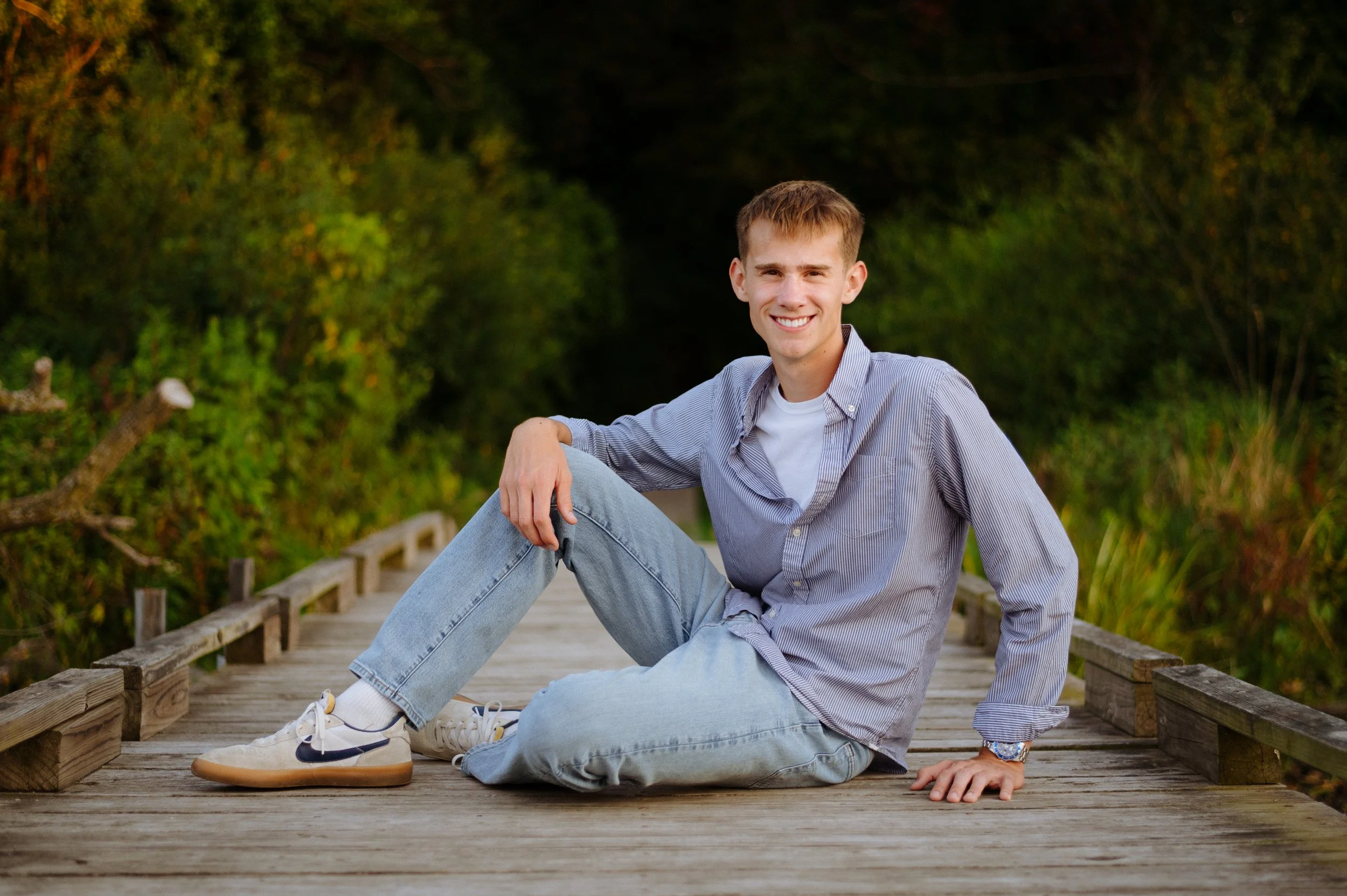A young man with short hair and a big smile sitting on a wooden bridge with green foliage in the background.