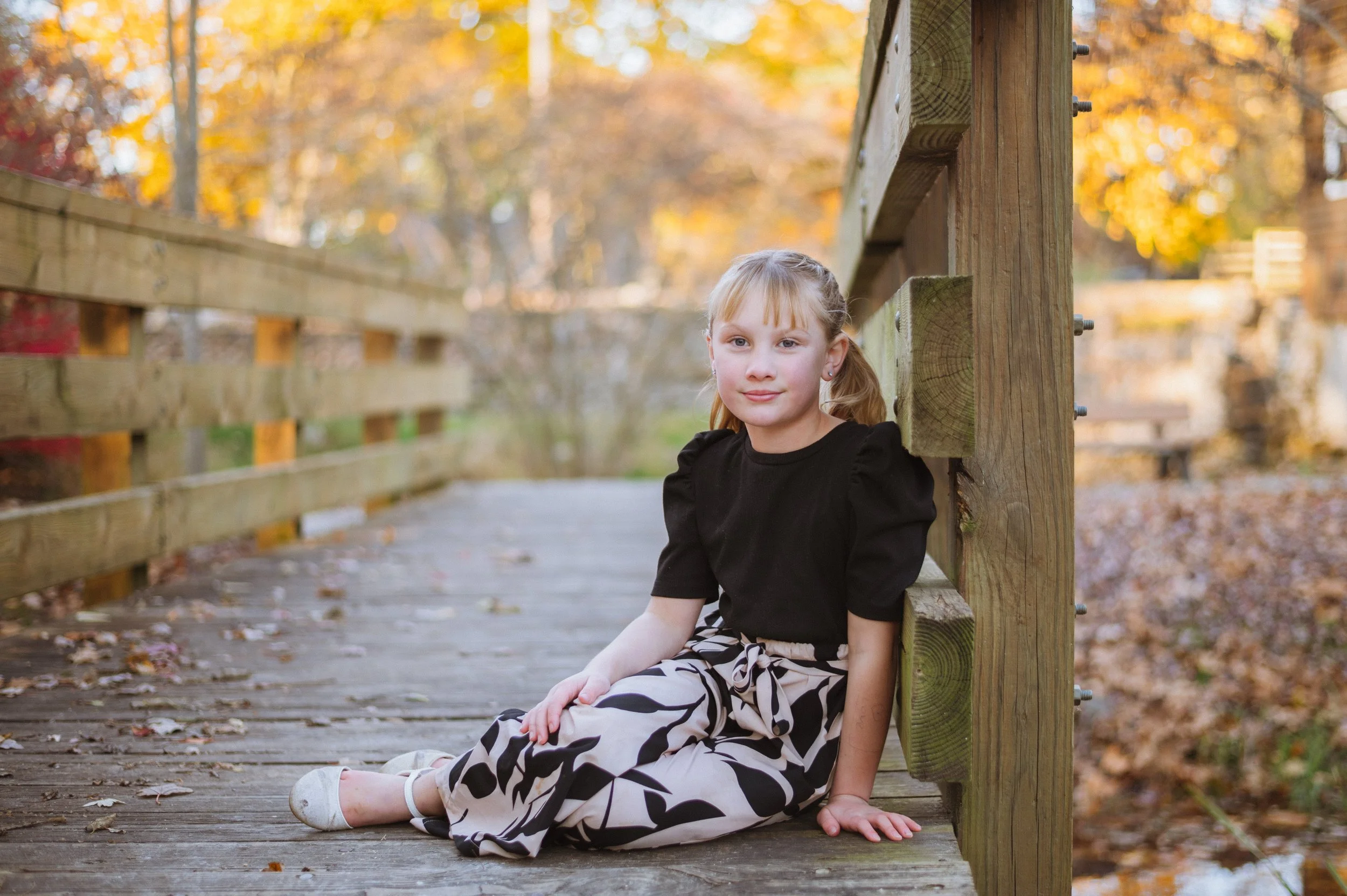 A young girl sitting on a wooden bridge outdoors during autumn, with fall leaves in the background.