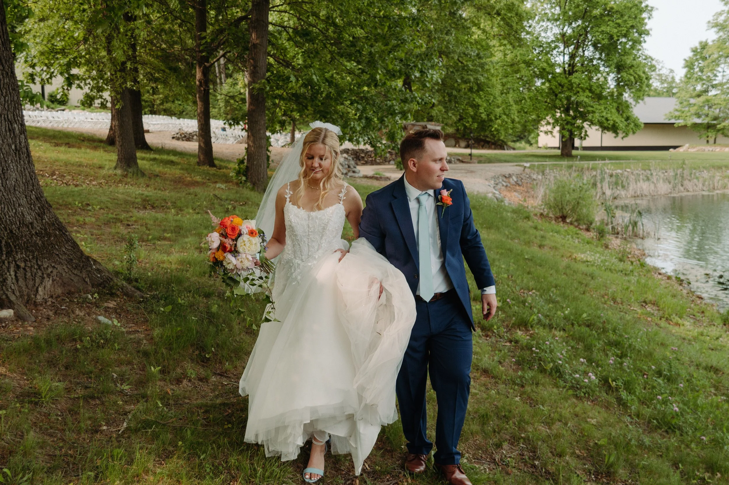 A bride and groom walking together outdoors near a lake, the bride holding a bouquet of flowers and lifting her dress slightly, surrounded by tall trees and green grass.