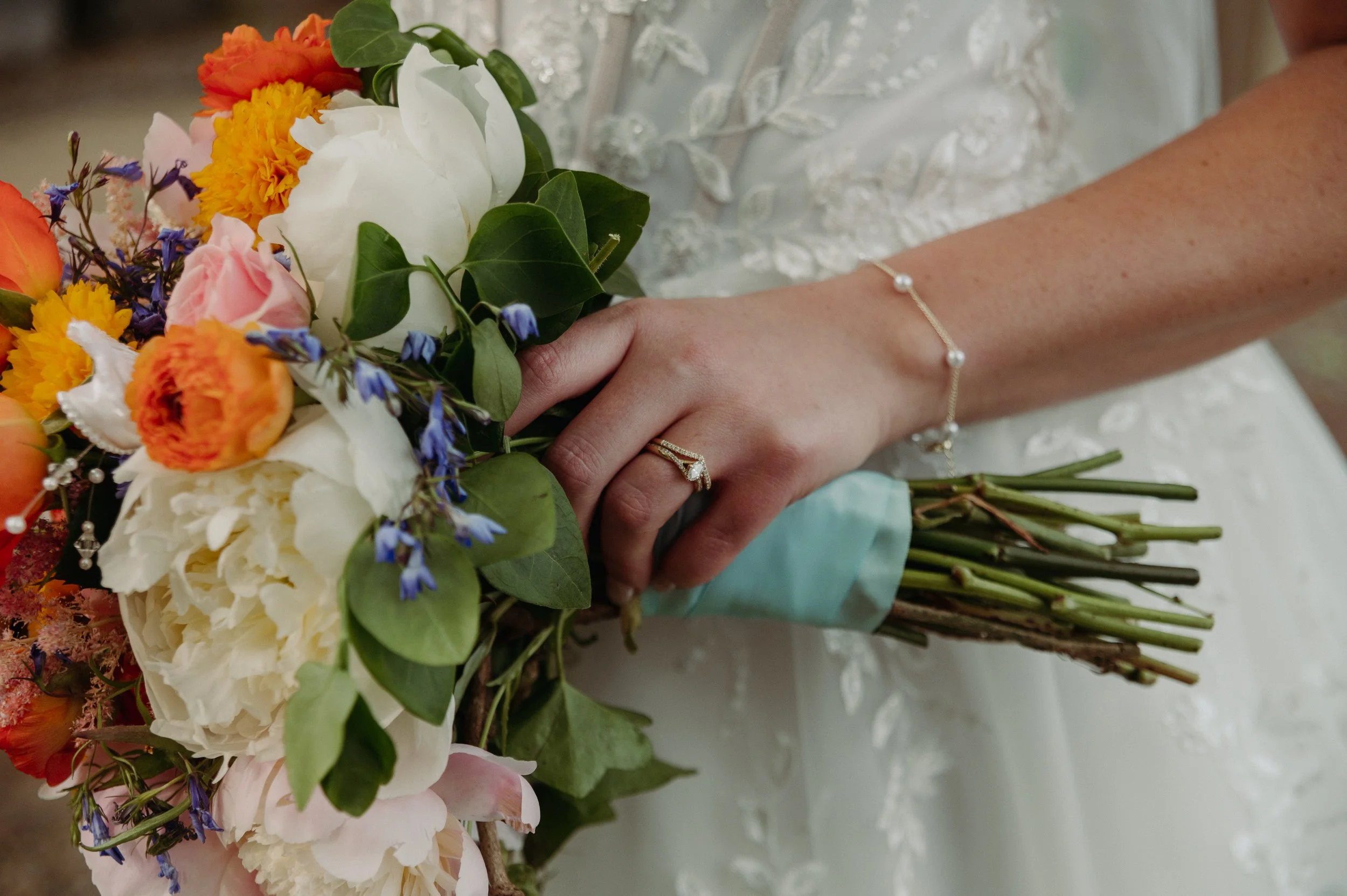 Close-up of a bride's hand holding a wedding bouquet with a diamond engagement ring and bracelet. The bouquet contains colorful flowers and green leaves.