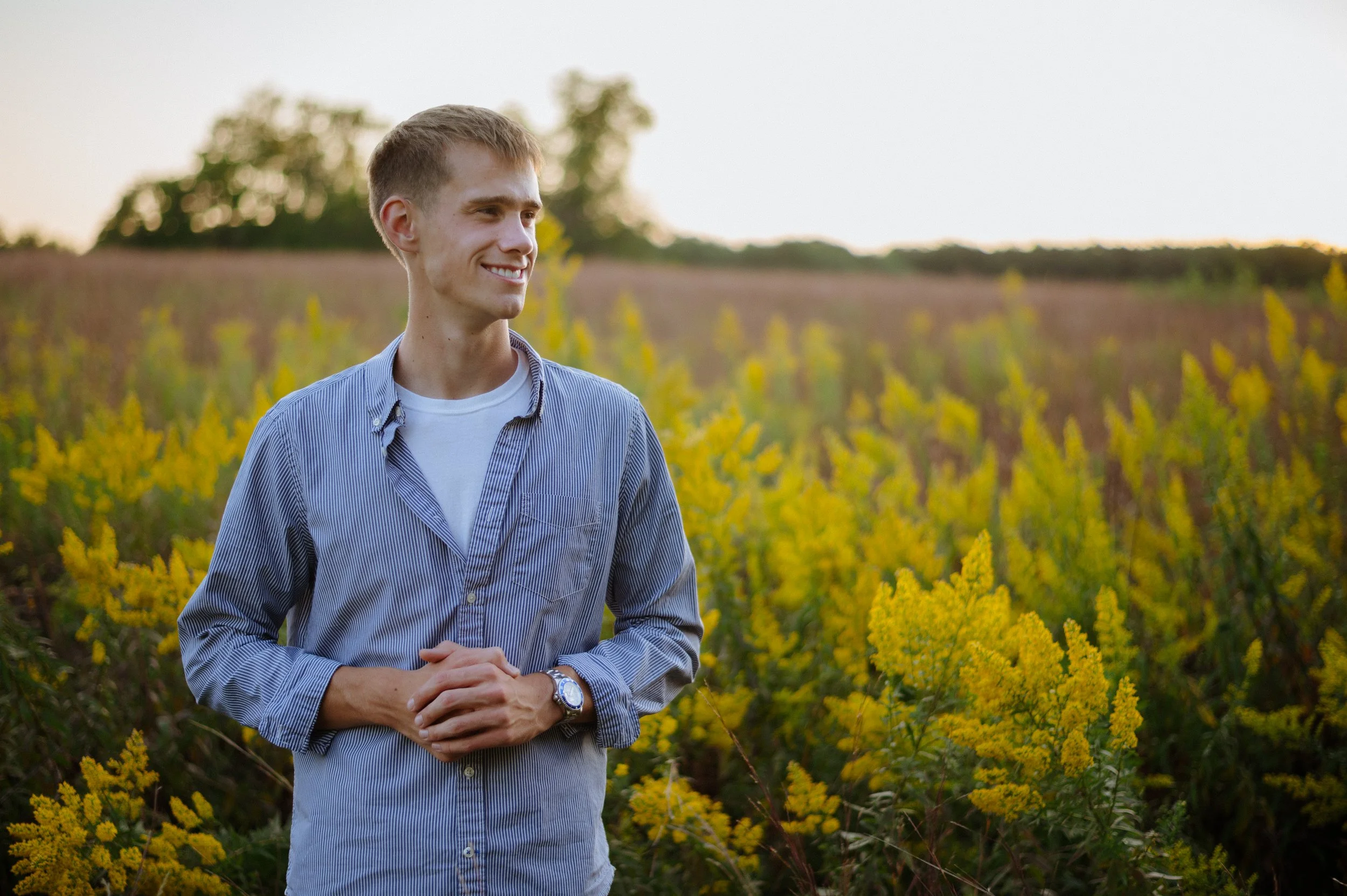 Young man standing in a field of yellow wildflowers during sunset, smiling and looking to the side.