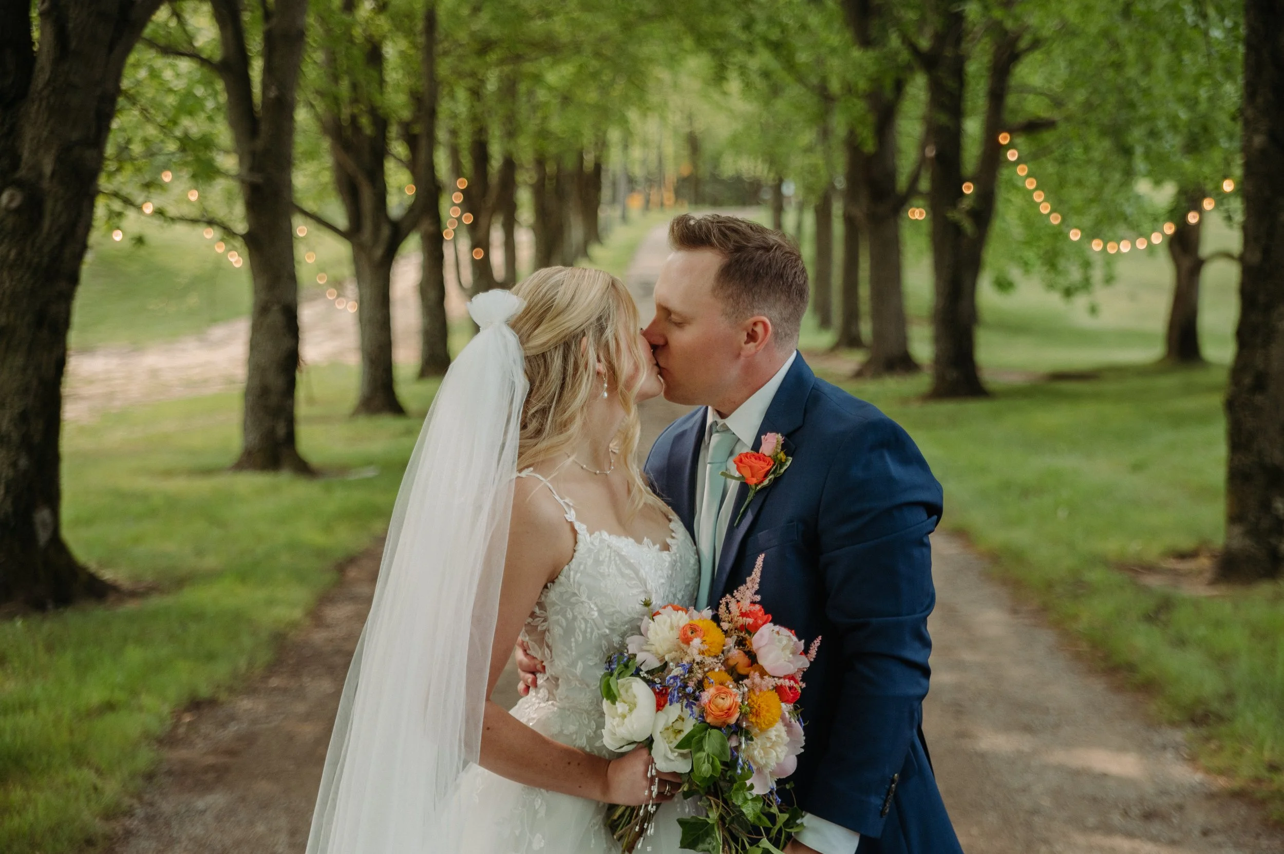 A newlywed couple kissing in a tree-lined outdoor setting with string lights hanging between trees.