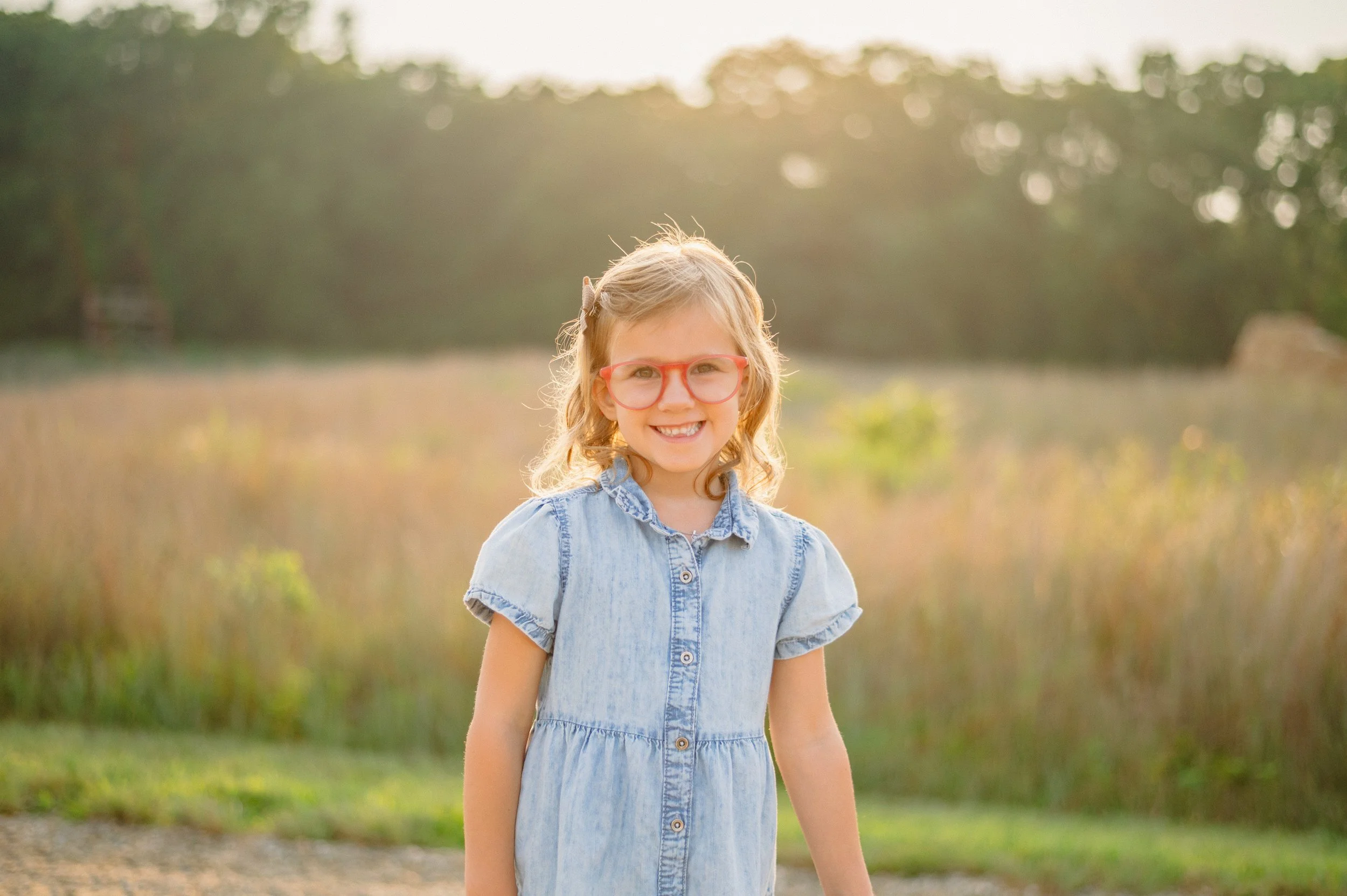A young girl with blonde hair and glasses is smiling outdoors at sunset, wearing a light blue denim dress.