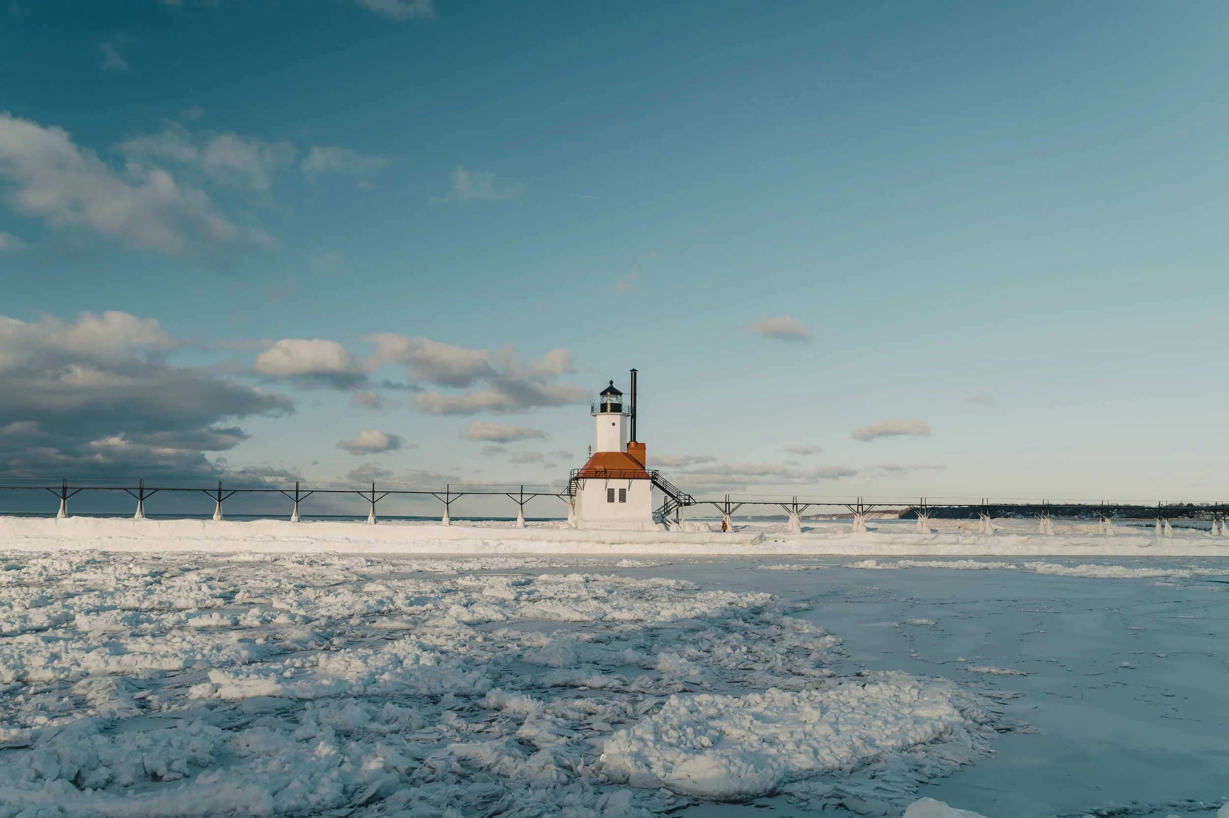 A lighthouse on a snowy, icy shoreline under a blue sky with scattered clouds.