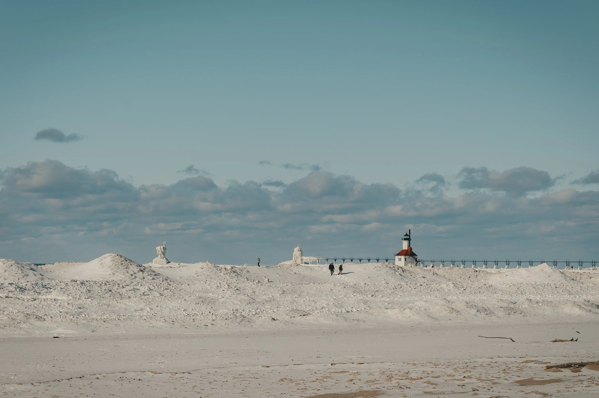 A lighthouse on a snowy shore with a wooden pier extending into the water, cloudy sky, and a few people walking.