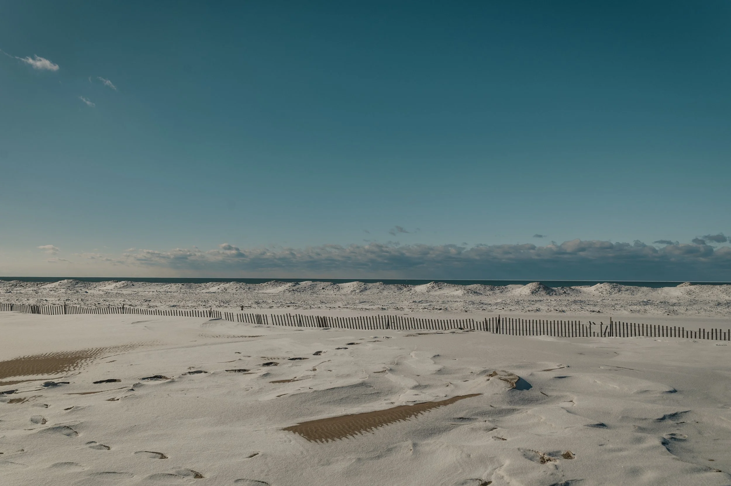 A sandy beach with footprints, a weathered wooden fence, sand dunes in the background, and a partly cloudy sky.