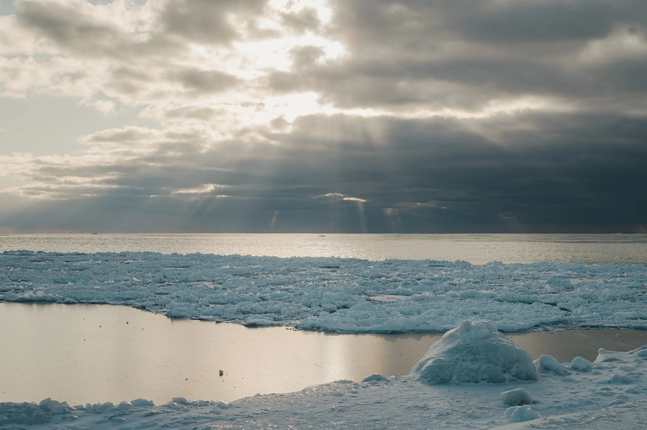 A cold, icy landscape with broken ice floating on water, overcast sky with sun rays breaking through clouds, and snow-covered ground.