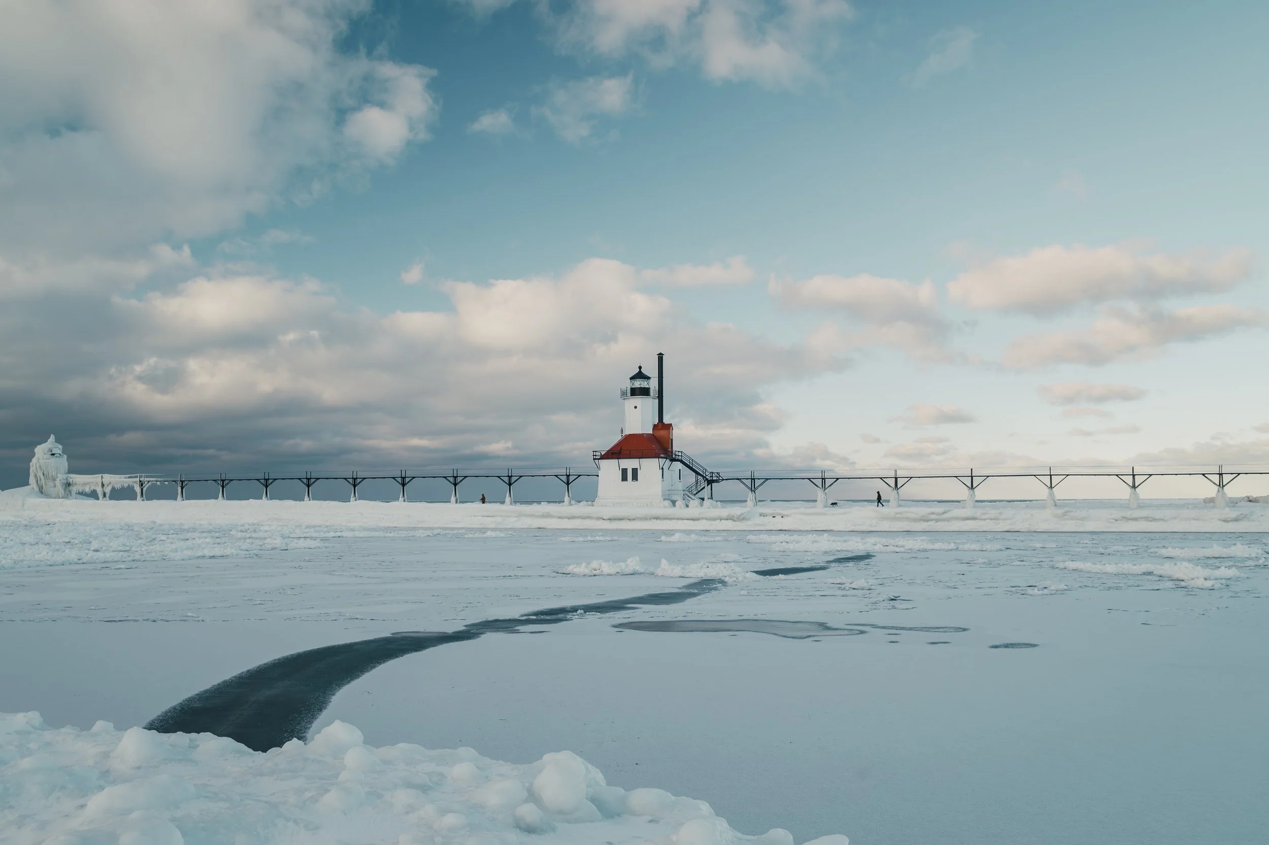A snow-covered landscape features a lighthouse with a red roof along a pier, extending into icy waters under a cloudy sky, with patches of ice and snow on the frozen surface.