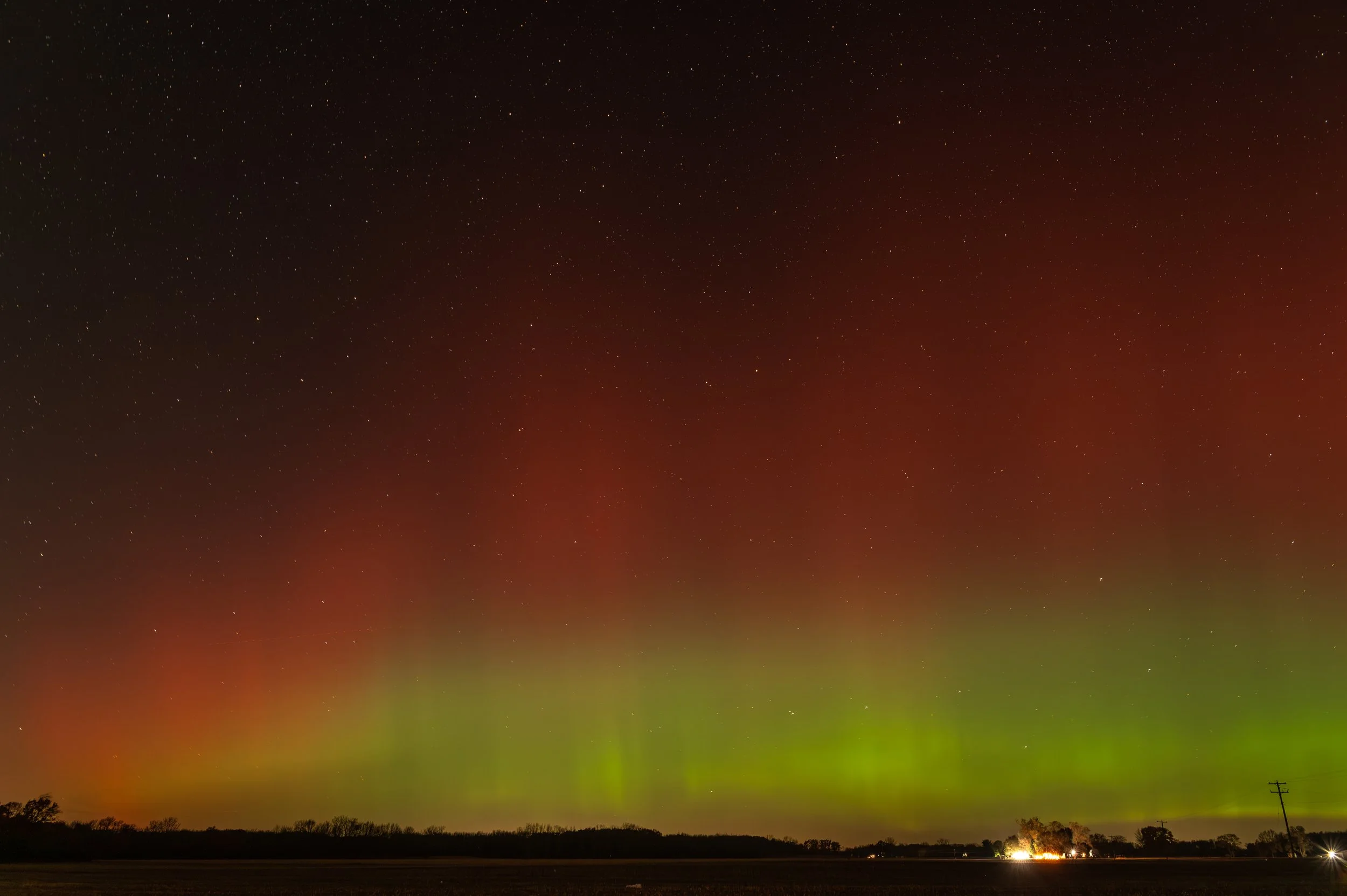 Night sky with visible stars and northern lights, with farm fields and trees at the horizon, and a couple of power poles on the right.