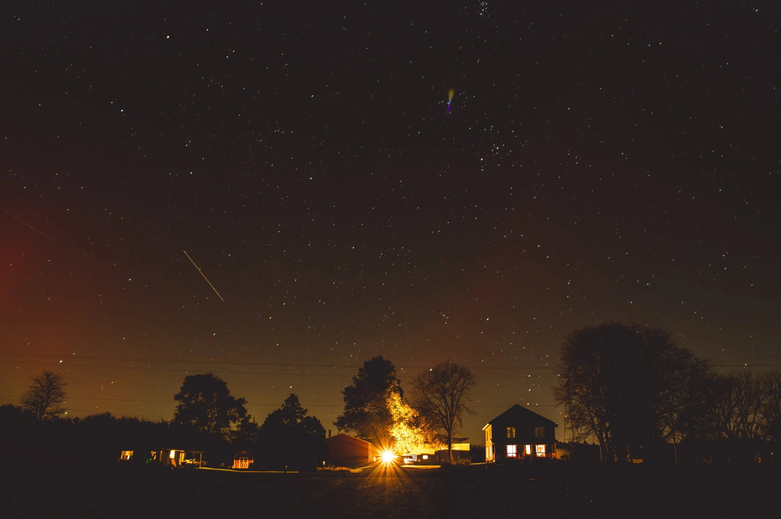 Nighttime scene of a rural residential area with several houses and trees silhouetted against a star-filled sky. A bright light emanates from a house in the foreground, and the sky shows many stars, a shooting star, and atmospheric haze near the hori