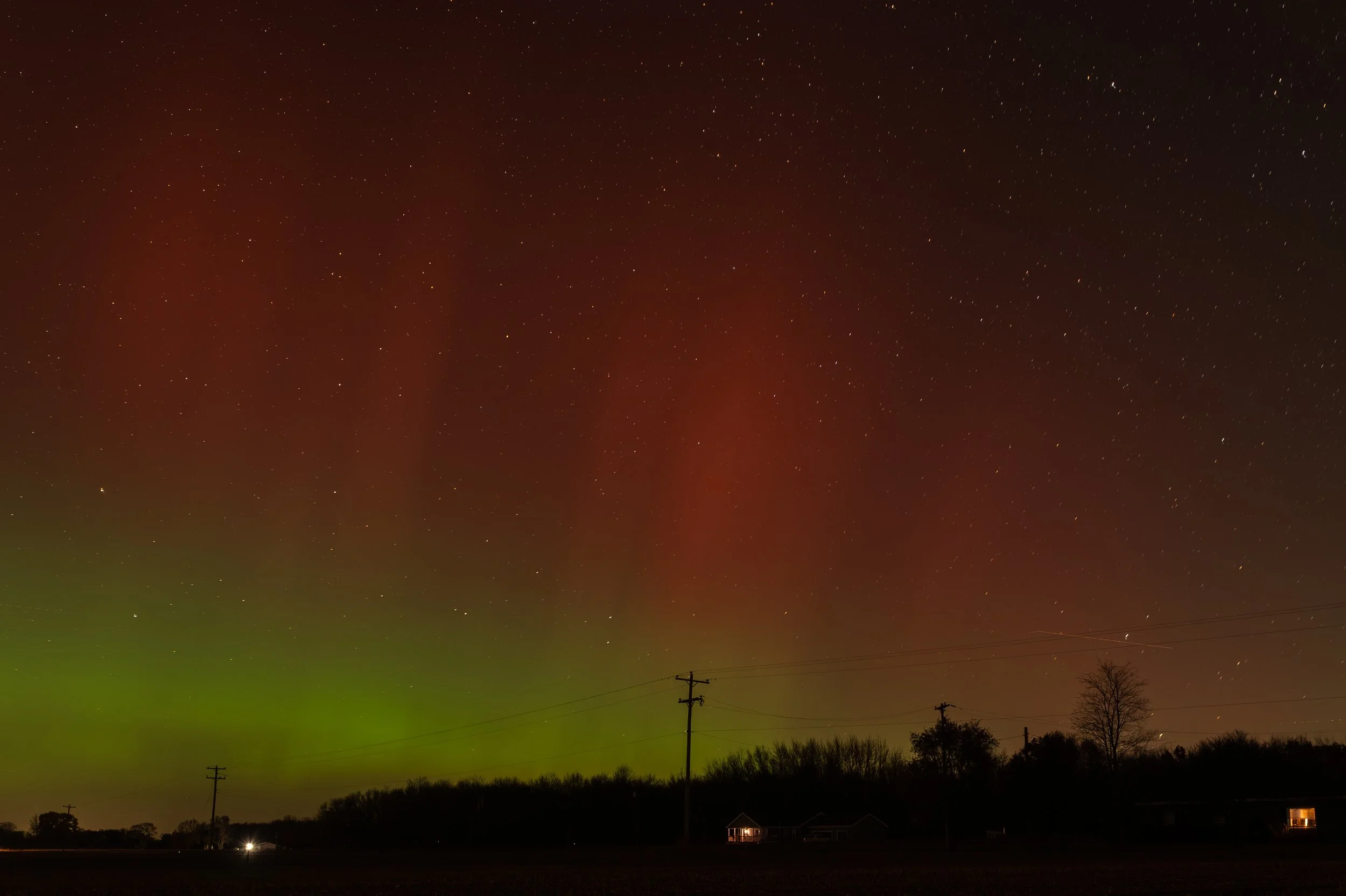 Night sky illuminated by the northern lights, with stars scattered across and silhouettes of houses, trees, and power lines at the ground.