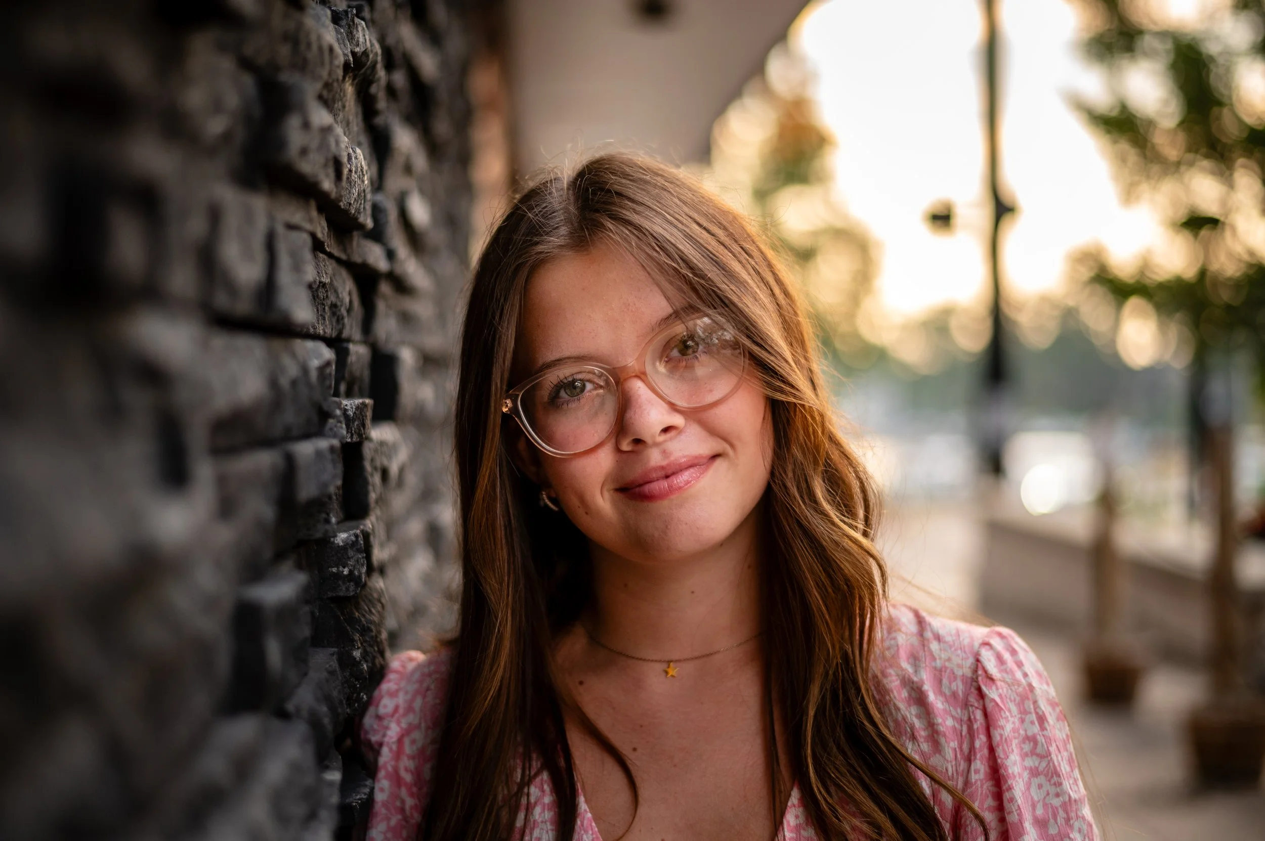 Young woman with long brown hair and glasses smiling outdoors in front of a stone wall, with a blurred background of trees and streetlights during sunset.