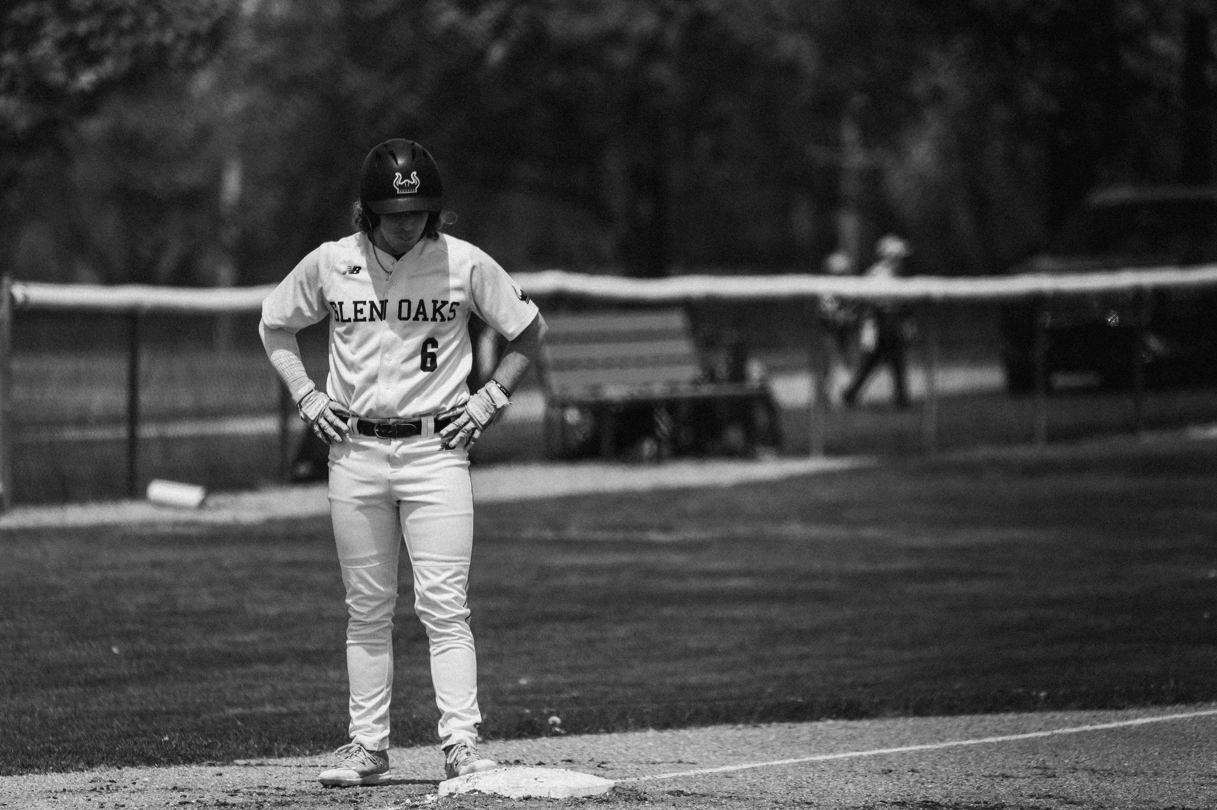 A baseball player standing on second base with hands on hips, wearing a uniform with 'Glen Oaks' and the number 6, a helmet, gloves, and cleats, on a baseball field with a fence and blurred figures in the background.