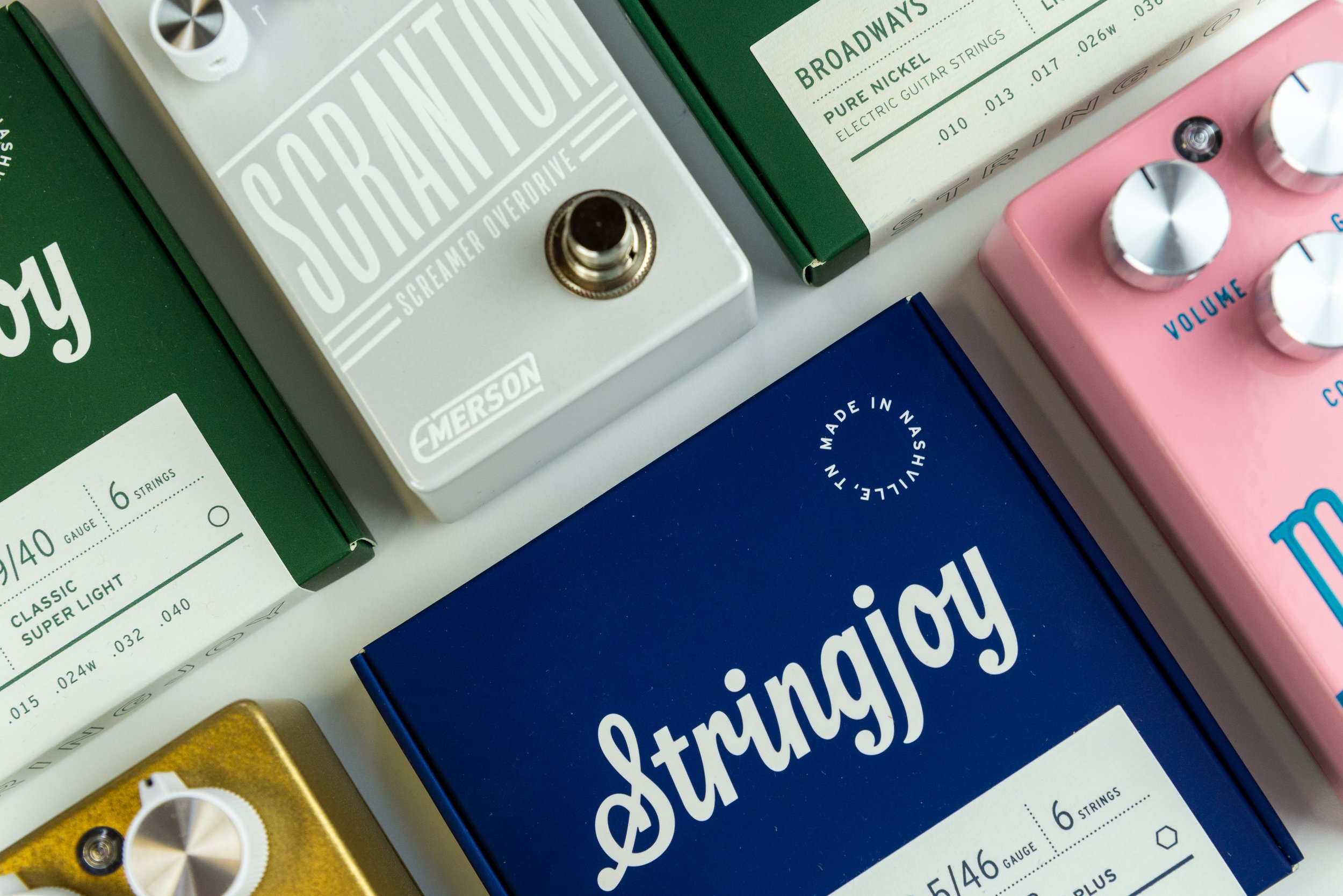 A collection of guitar effect pedal boxes and a pink guitar pedal on a white surface. The pedal boxes are green, white, yellow, and blue, each labeled with brand and type, with control knobs visible on some.