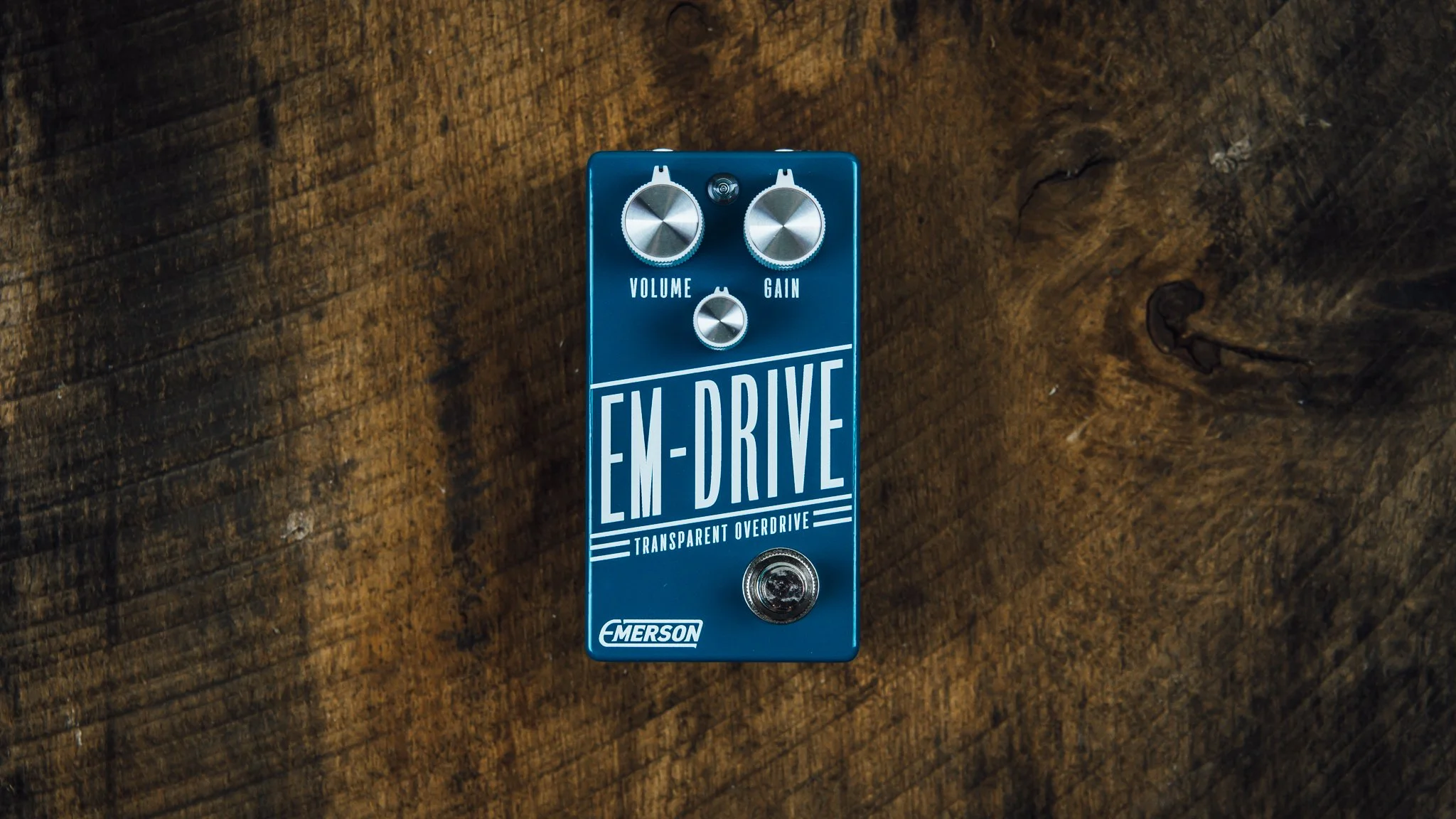 Blue guitar effects pedal labeled 'EMERSON EM-DRIVE' with controls for volume, gain, and a small footswitch, placed on a wooden surface.