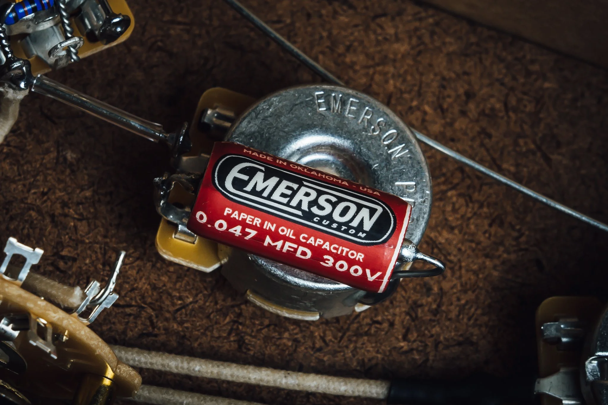 Vintage Emerson paper oil capacitor with red and black label, lying on a brown textured surface, surrounded by electronic components and wires.