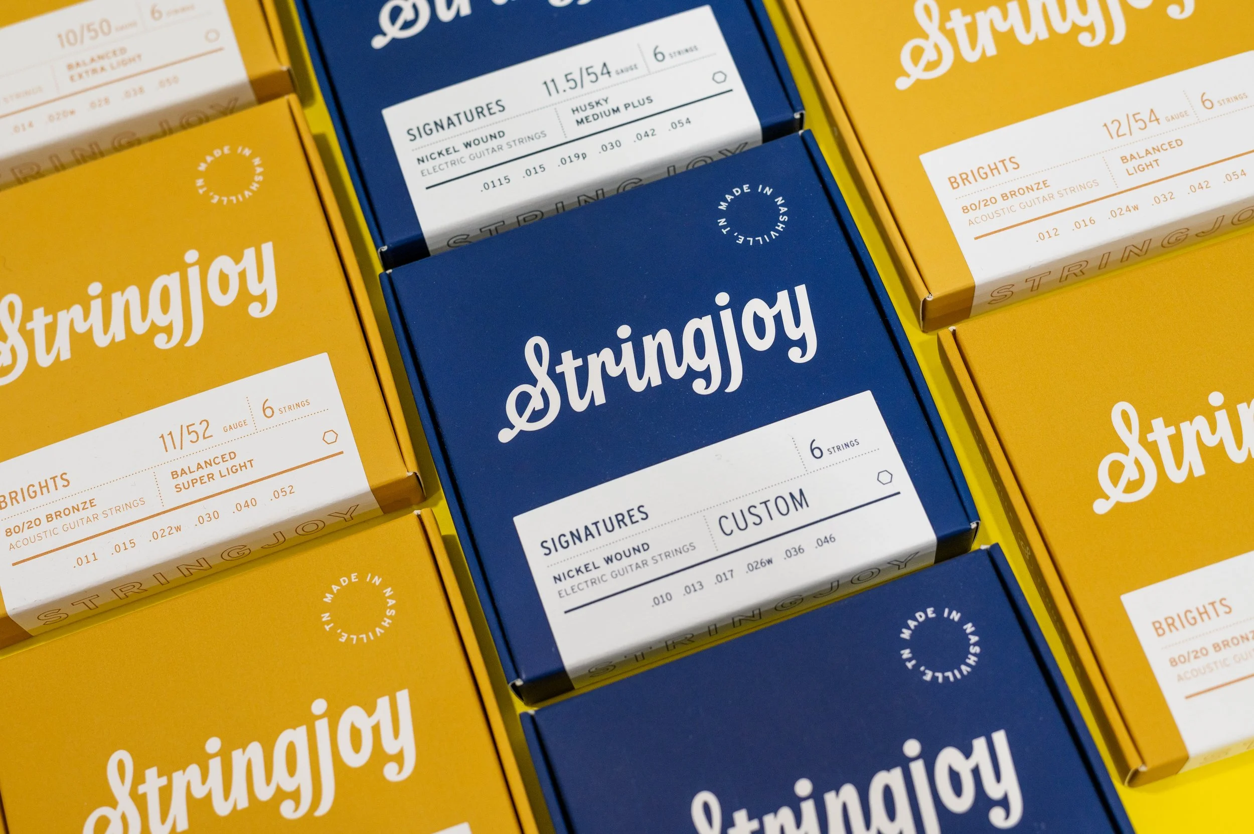 Multiple boxes of Stringjoy guitar strings in yellow and blue packaging, featuring brand name and string specifications.