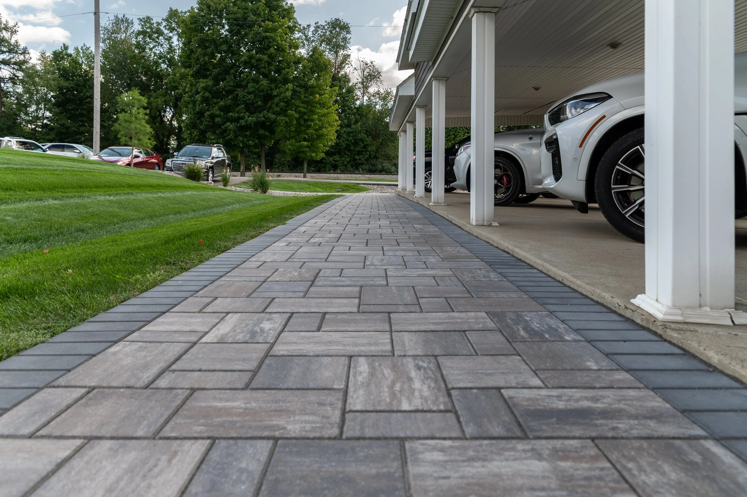 A sidewalk running past a grassy lawn and a covered parking area with white and silver cars, with trees and a cloudy sky in the background.