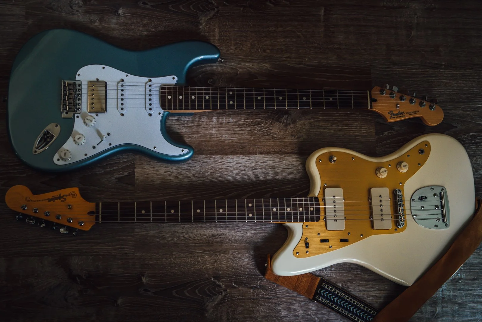 Two electric guitars lying on a wooden floor, one blue with a white pickguard and the other white with yellow pickguard, both with multiple control knobs and pickups.