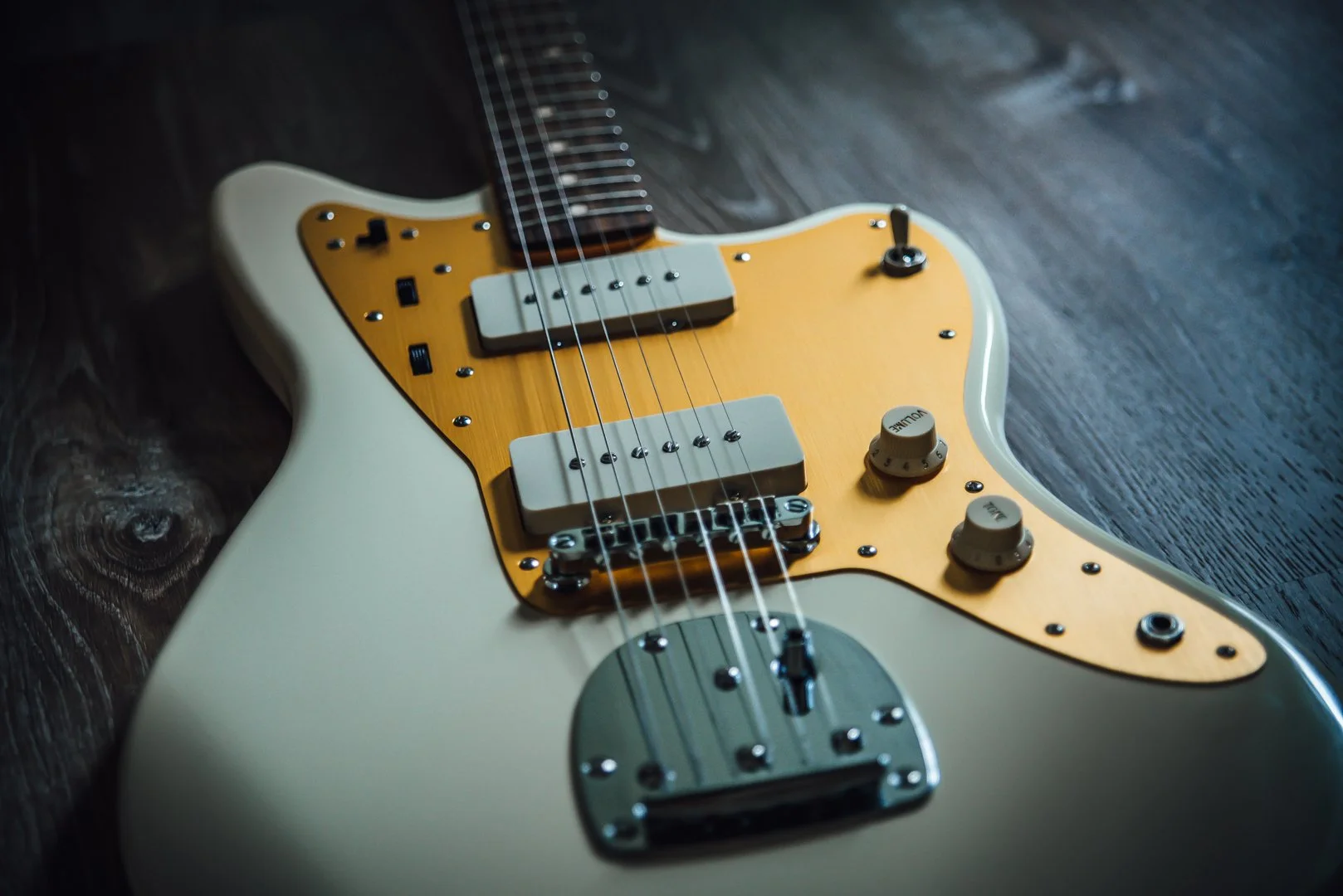 Close-up of a cream-colored electric guitar with a yellow pickguard, lying on a dark wooden surface. Squier JMascis