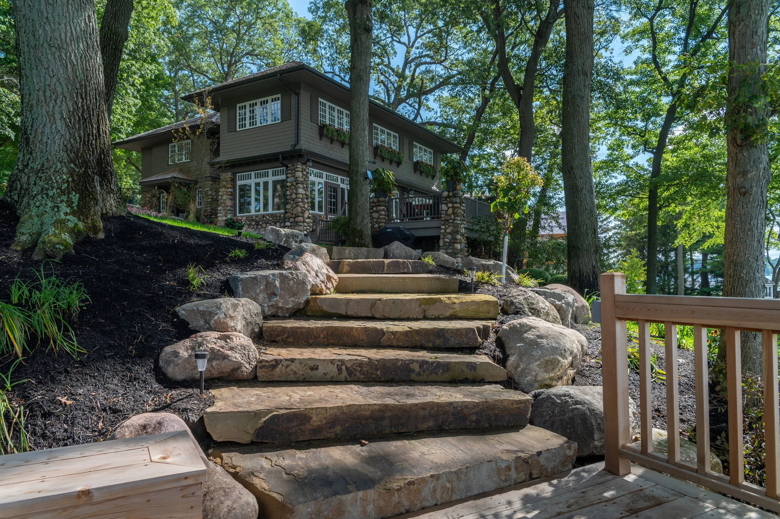 Stone steps leading up to a house in a wooded backyard, with a wooden deck and large trees surrounding the area.