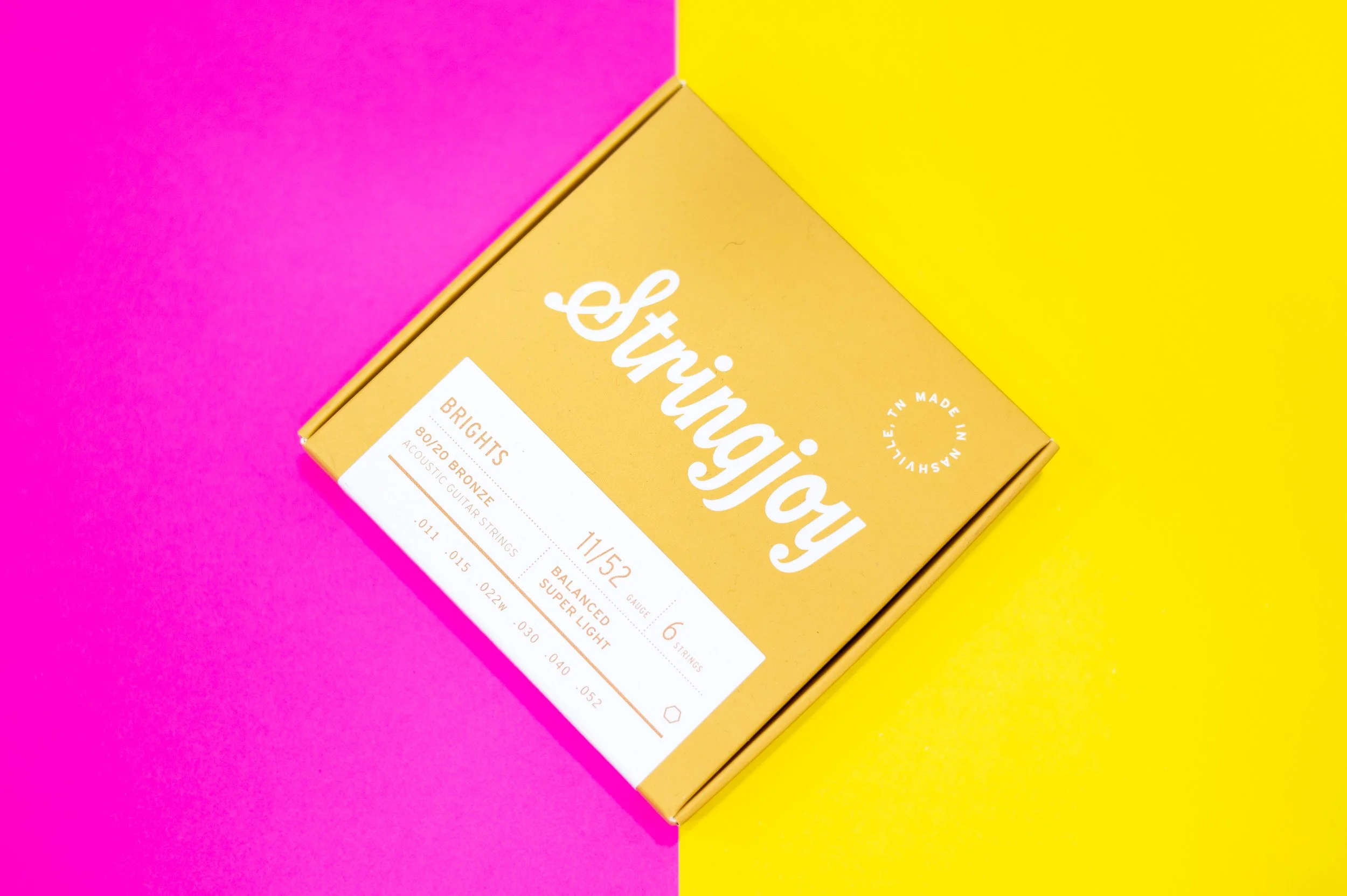 A yellow box of Strings & Things guitar strings resting on a pink and yellow split background.
