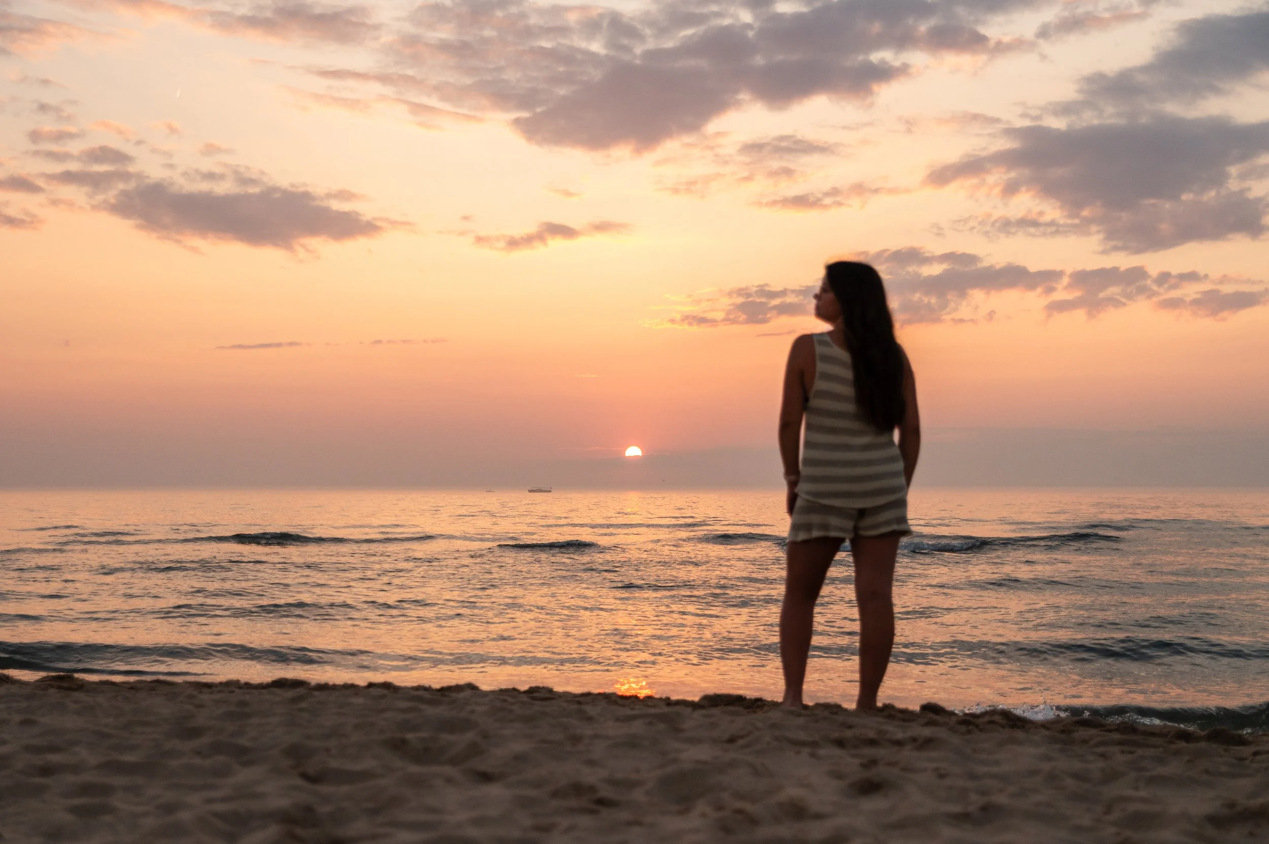 A woman stands on the sandy beach, looking out at the sunset over the ocean with a partly cloudy sky.