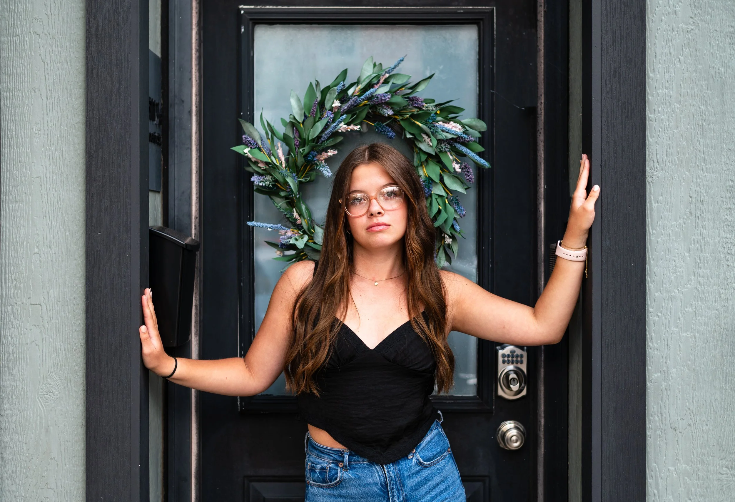 Young woman standing at a black front door with a decorative floral wreath. She has long brown hair, glasses, wearing a black sleeveless top and blue jeans.