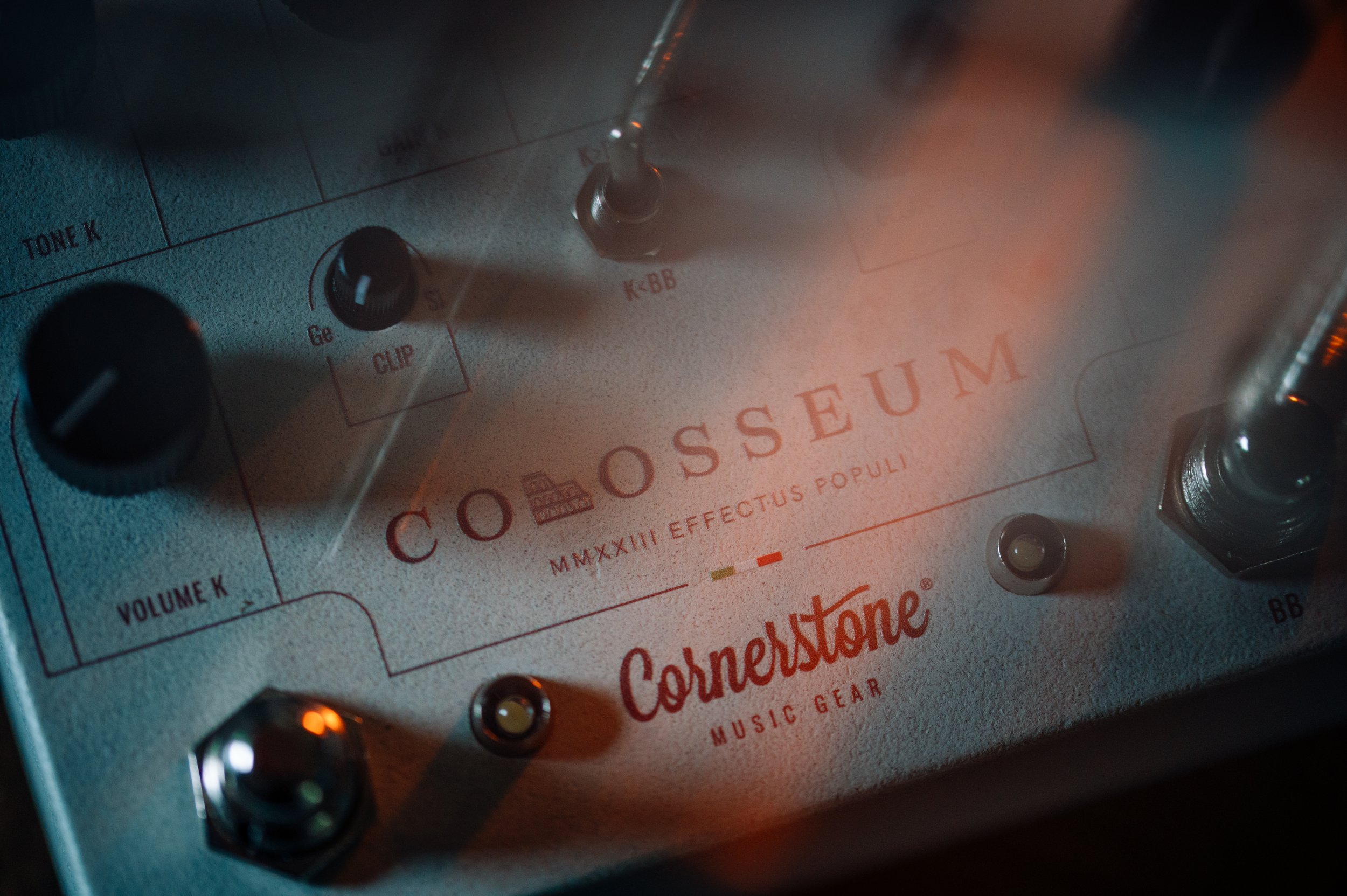 Close-up of a music gear effects pedal labeled 'Colosseum' by Cornerstone, with various control knobs and switches, illuminated by dim lighting.