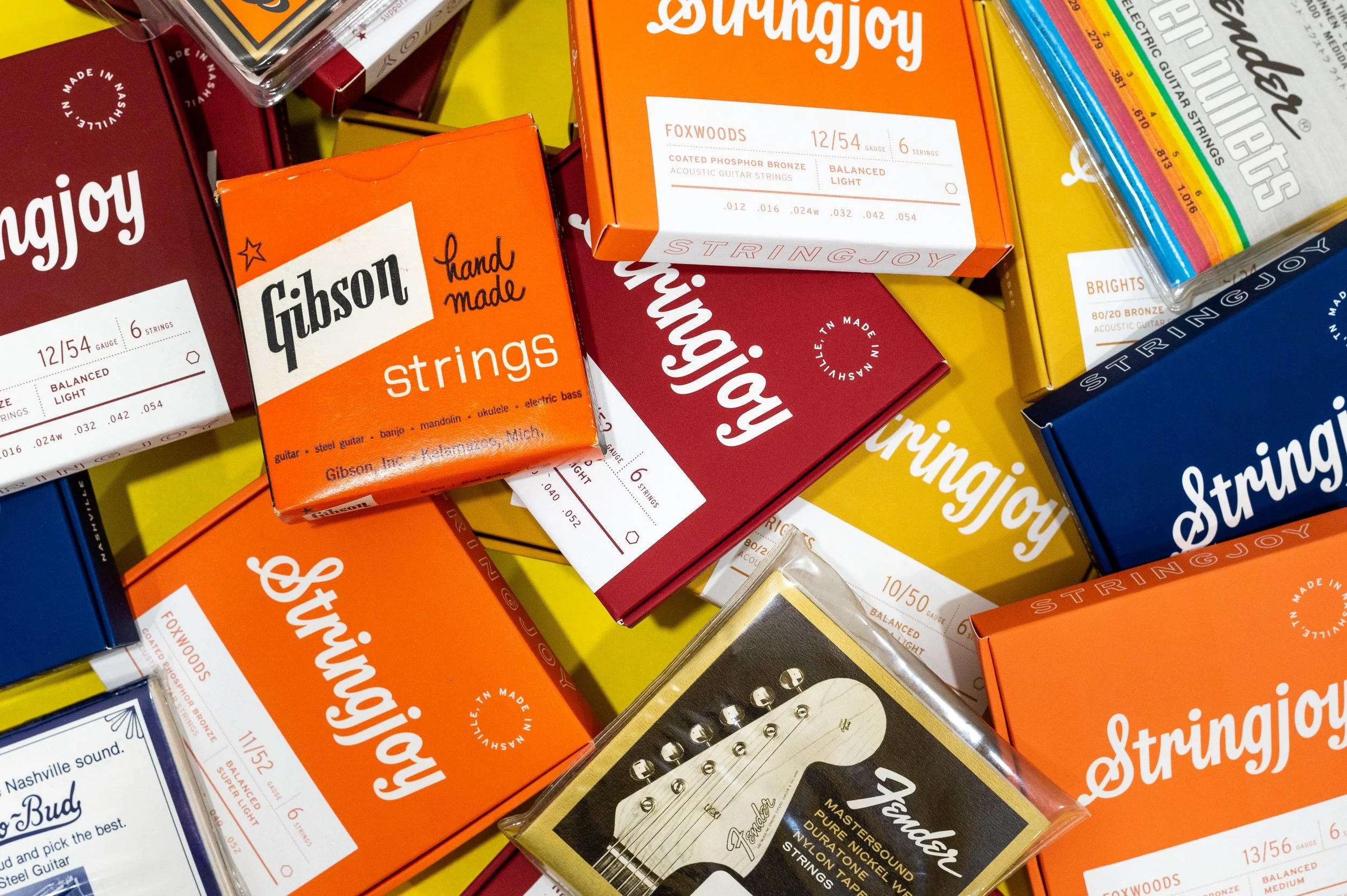 A collection of colorful boxes of guitar strings from brands like Gibson, Fender, and Ernie Ball, spread out on a bright yellow surface.