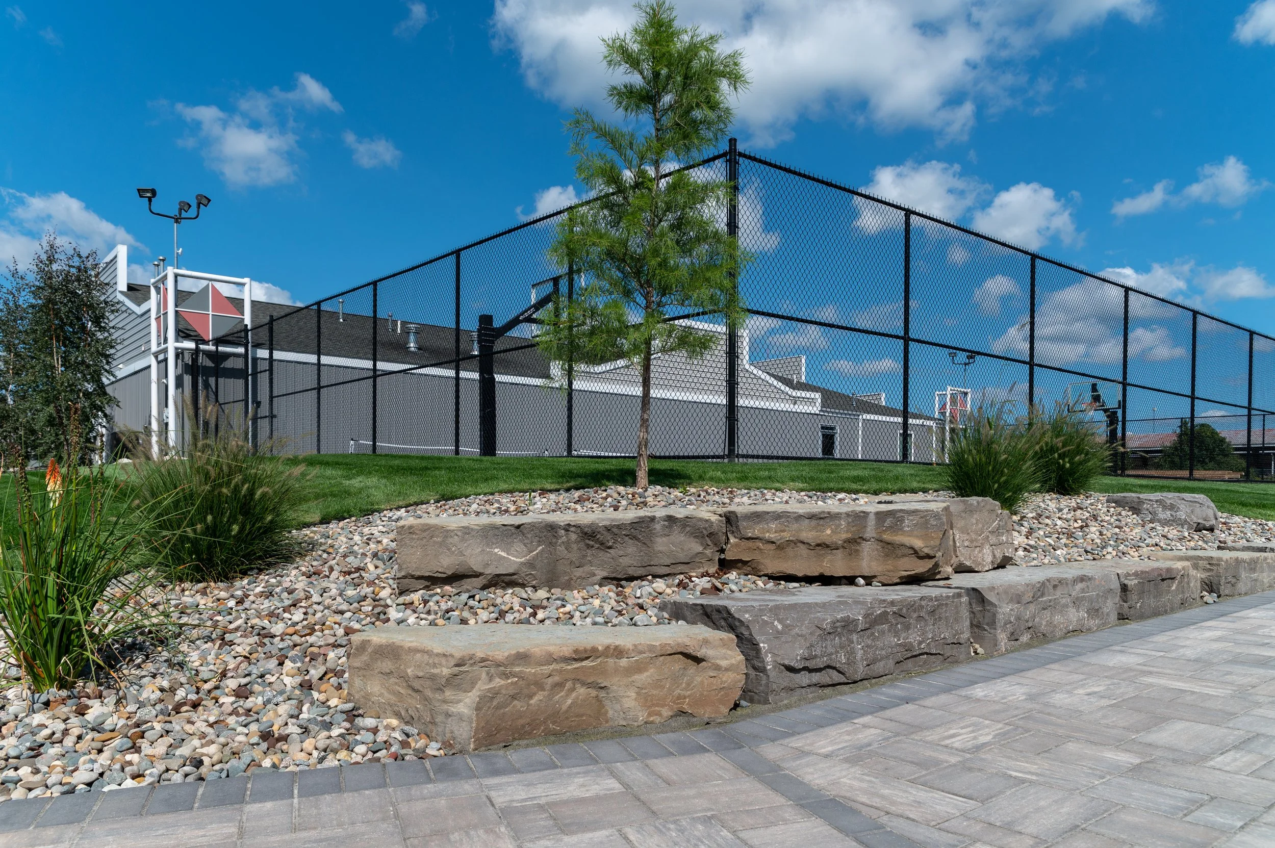 A landscaped area with a small green tree, decorative rocks, and large stone steps in front of a black chain-link fence and a building with grey walls and a black roof, under a blue sky with scattered white clouds.
