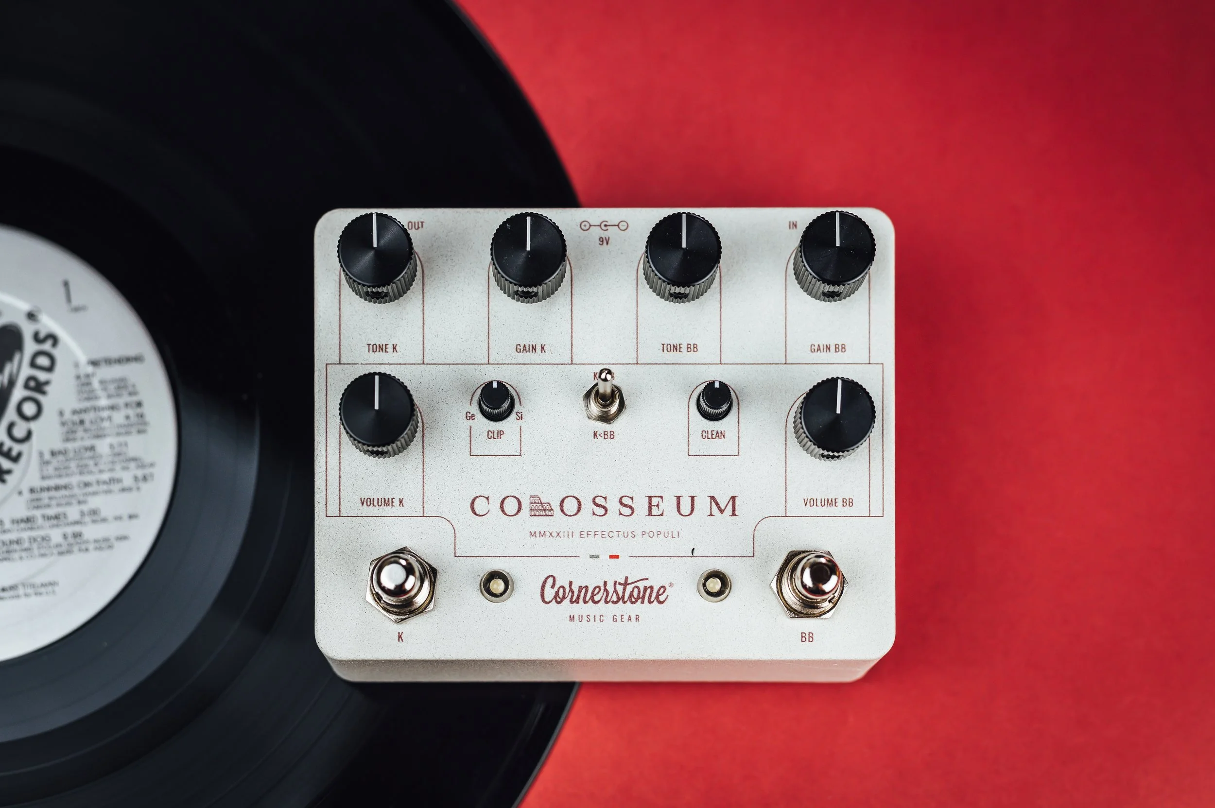 A musical effects pedal labeled 'Colosseum' by Cornerstone Music Gear, with various knobs and switches, placed next to a vinyl record on a red surface.