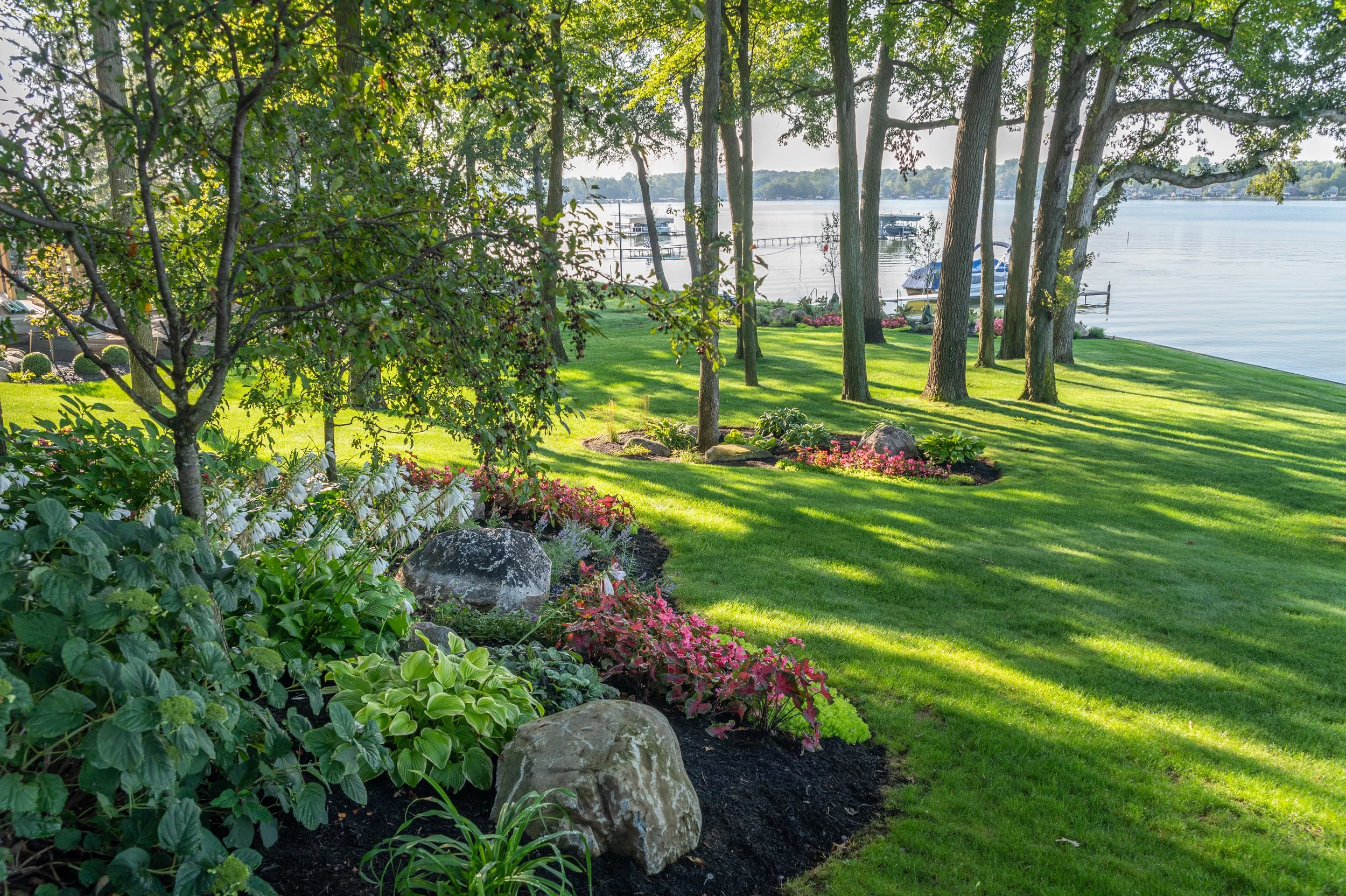 A scenic lakeside garden with lush green grass, trees, flowering plants, and rocks. The background shows a calm lake with boats docked and a clear sky.