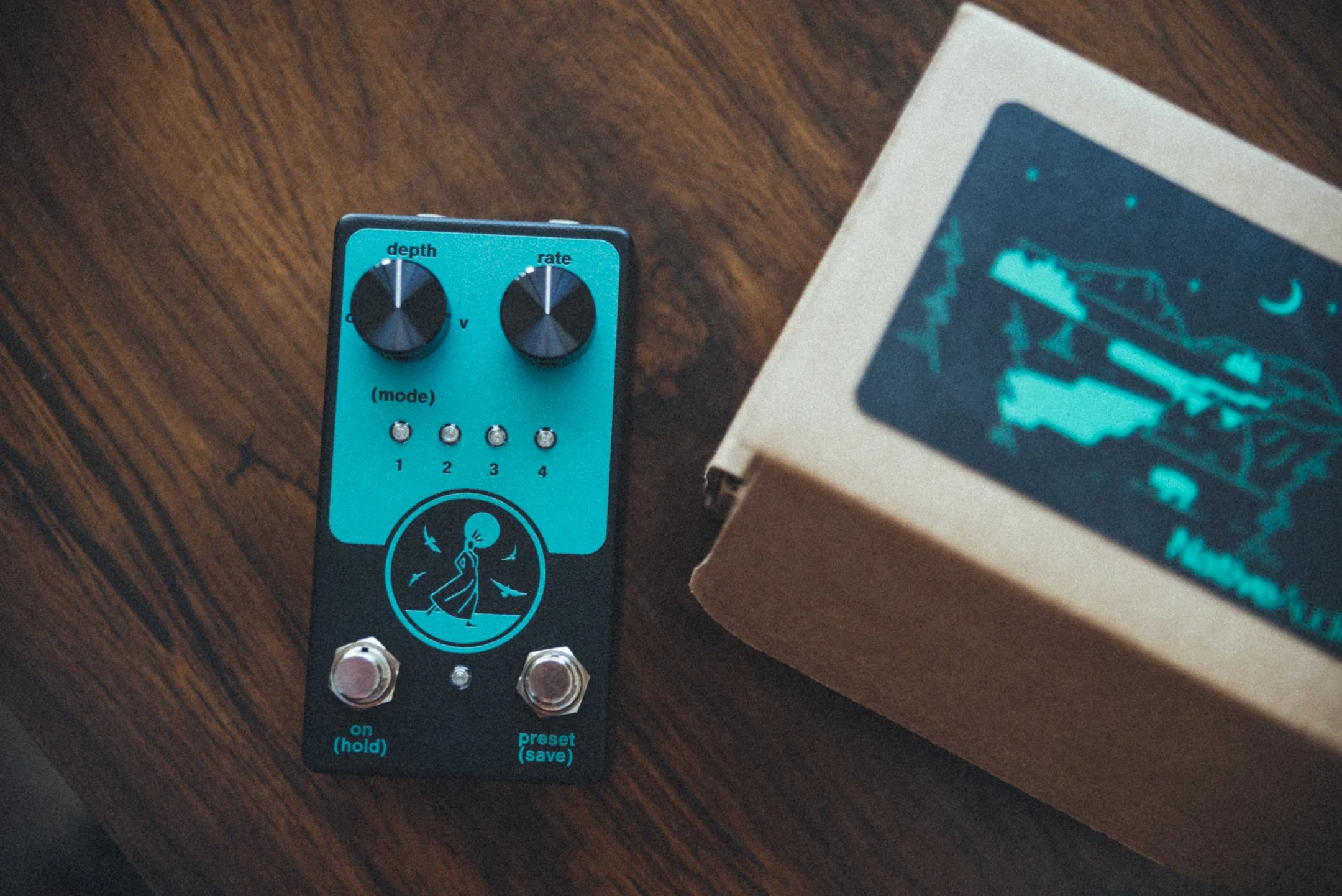 A guitar effects pedal with a blue and black design, featuring knobs labeled 'depth' and 'rate', four mode indicator lights, an illustration of a girl and birds, and two footswitches, placed on a wooden surface next to a cardboard box with blue graph