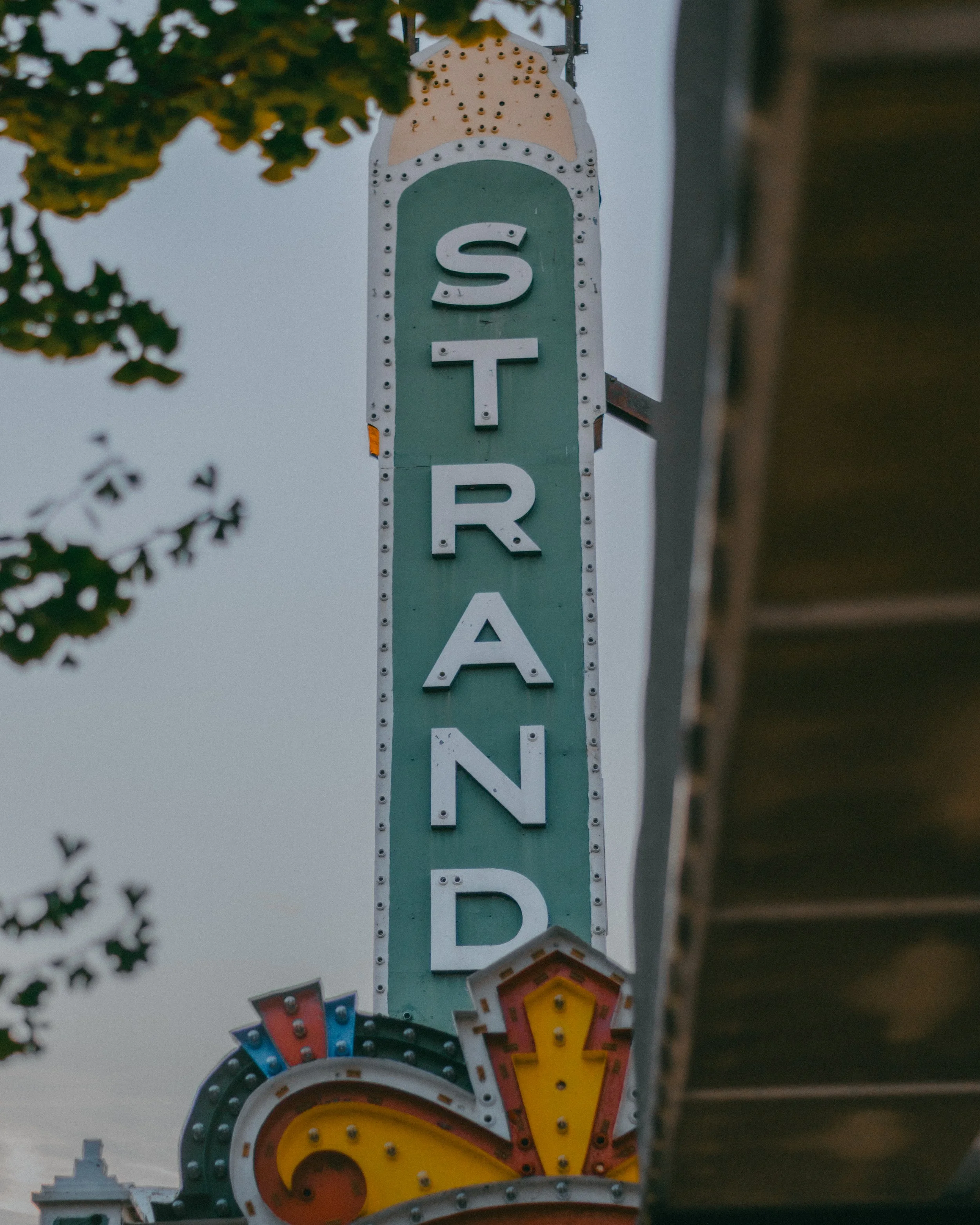 Vertical neon sign spelling 'STRAND' with a decorative arrow or design at the bottom and a small arrow pointing downward.