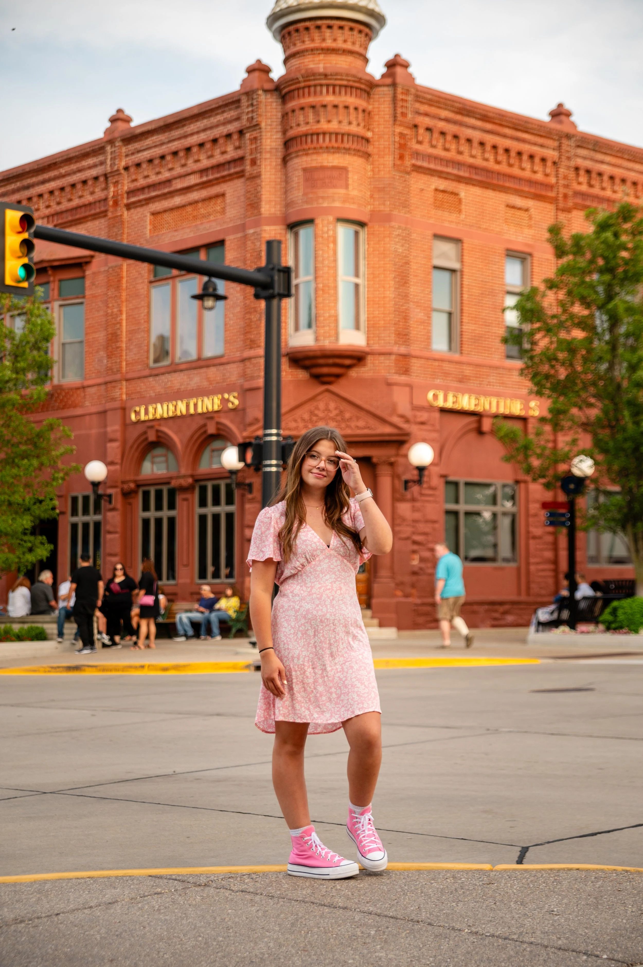 A young woman in a pink dress and pink high-top sneakers standing on a city street at sunset, with a red brick building named "Clementine's" in the background.