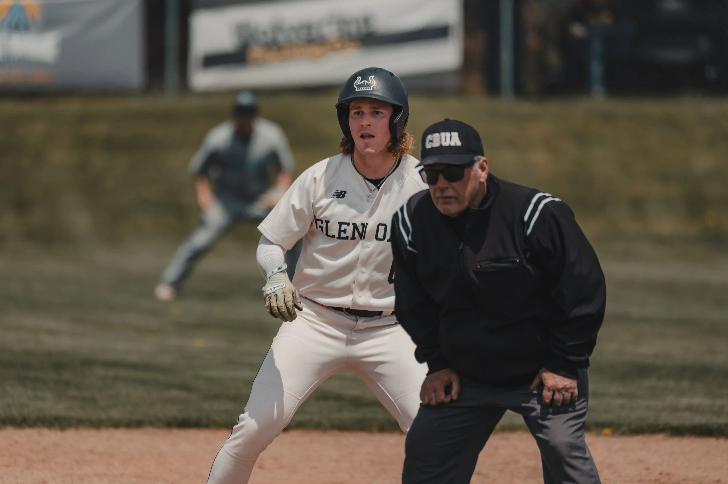 A baseball player in a white uniform with 'Glen' on the front is looking ahead, wearing a helmet and batting gloves, on a baseball field during a game. An umpire wearing a black jacket, cap, and sunglasses is in front of him, leaning forward. Another
