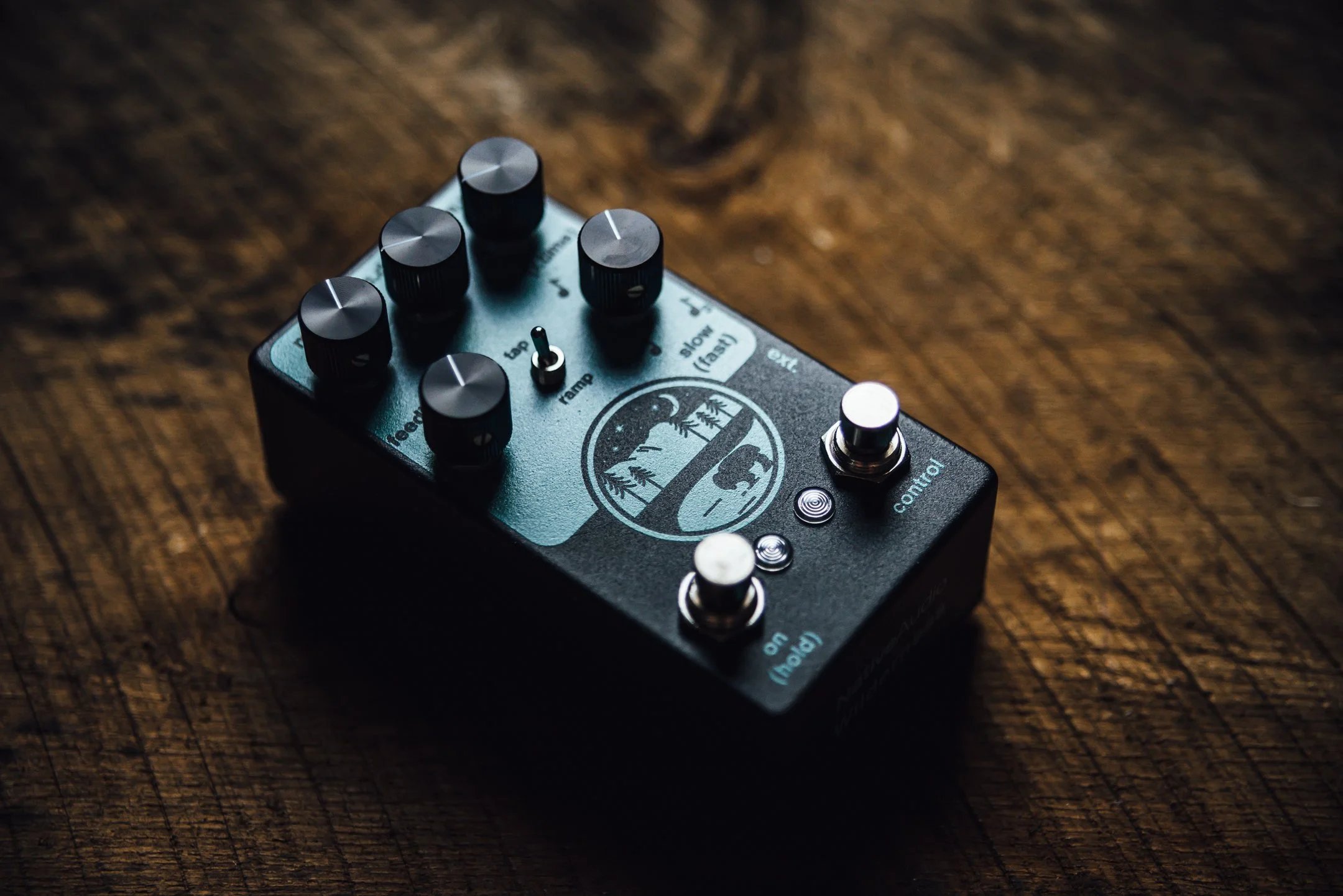 A black guitar effects pedal with six control knobs and two foot switches, resting on a wooden surface. The pedal has a graphic of a mountain landscape with a bear and trees.