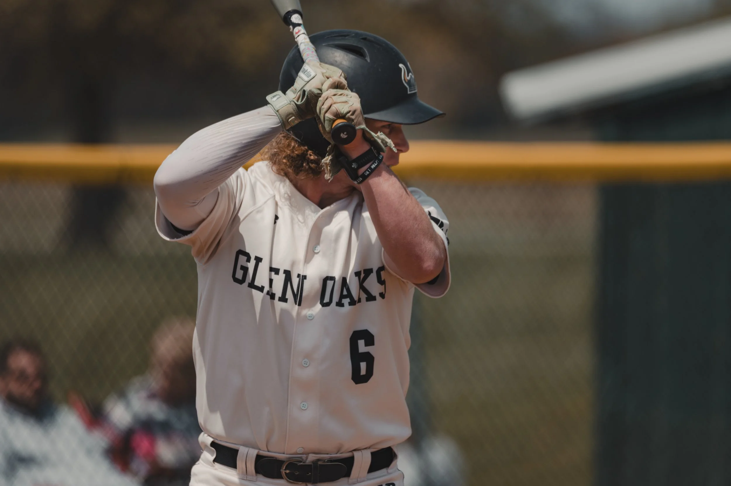 A baseball player in a white uniform with 'GLEN OAKS' and the number 6, standing at bat with a helmet and batting gloves, holding a bat up ready to swing.