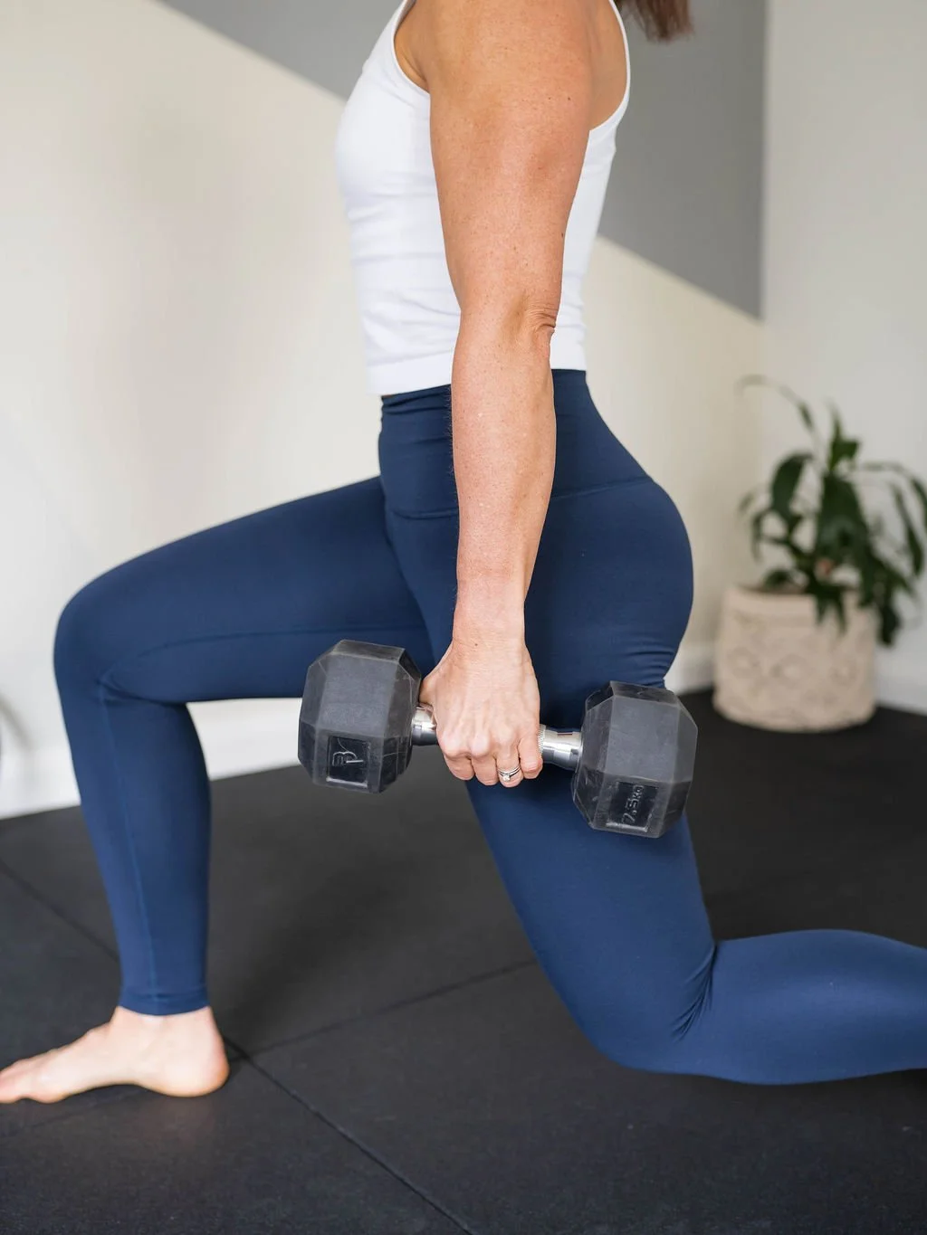 Woman doing a lunge exercise indoors, holding a dumbbell in her right hand, wearing a white tank top and dark blue leggings.