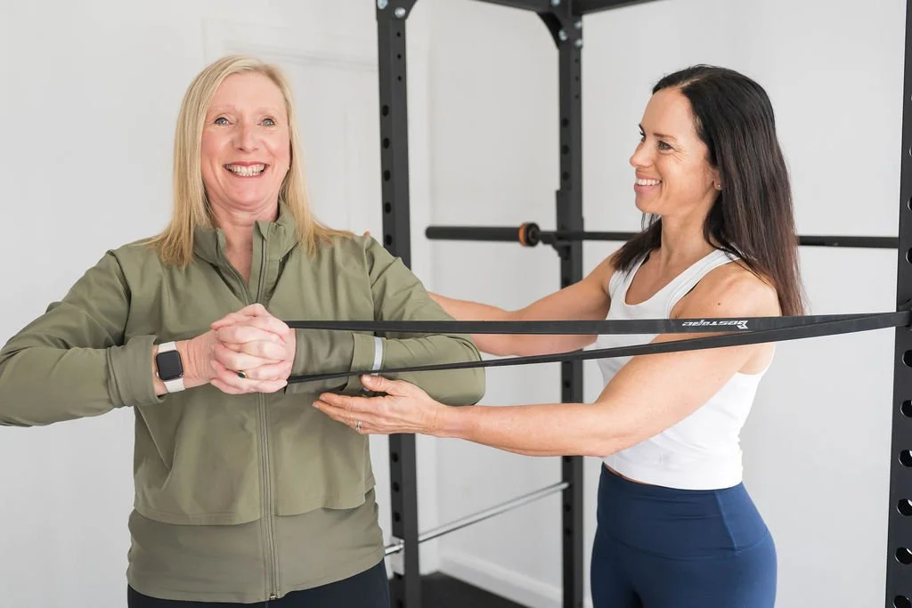 Two women exercising with resistance bands in a home gym, smiling and standing near a black metal workout rack.