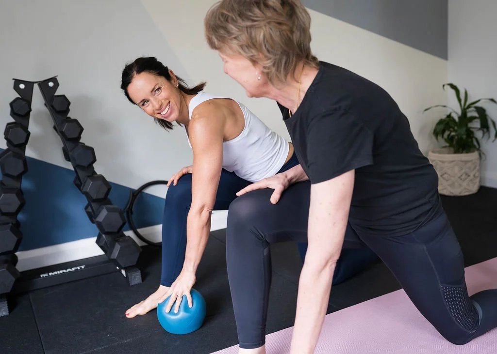 Woman performing a kettlebell exercise with assistance from a trainer in a gym.
